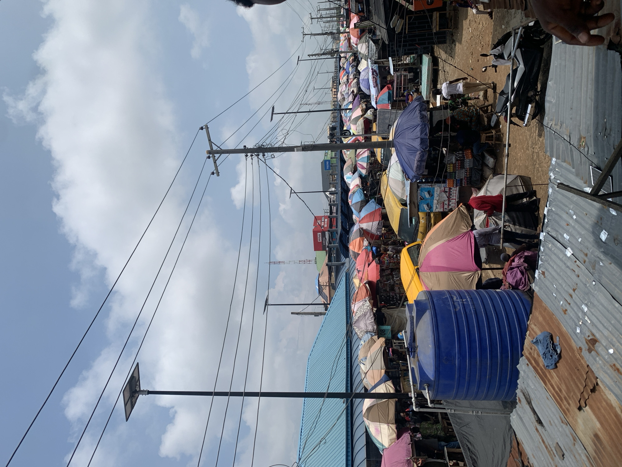 This is a street scene in Ikorodu, Nigeria, likely a market. The image is dominated by a multitude of colorful umbrellas providing shade for market stalls and goods. People are visible, engaged in shopping or selling, though their faces are not clearly discernible due to the distance and the nature of the scene. Objects include market wares, large blue water tanks, electrical poles with numerous wires, a motorcycle, and corrugated metal roofing. The sky is partly cloudy, suggesting it is daytime. The scene appears bustling and active, typical of a busy marketplace. There is some visible text on signs, including "TECNO" and "INFINIX", which are mobile phone brands, indicating these might be electronics stalls. The overall impression is one of a lively and crowded market environment under a cloudy sky.