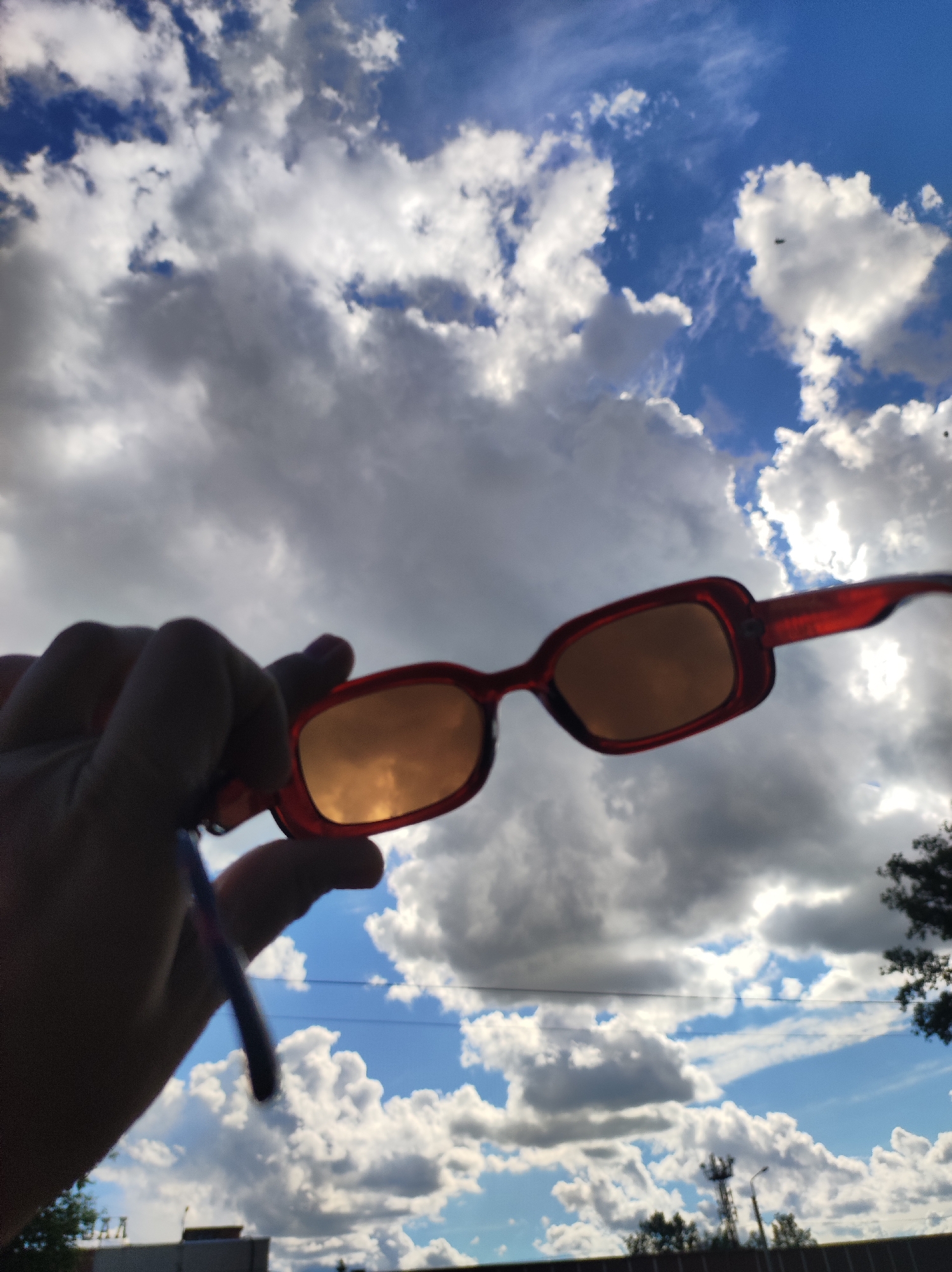 A hand is holding a pair of red-framed sunglasses against a backdrop of clouds and blue sky. The sunglasses lenses reflect the sky and clouds. In the lower portion of the image, a building, foliage, and a utility pole are visible. The location is Chaikovsky, Russia.