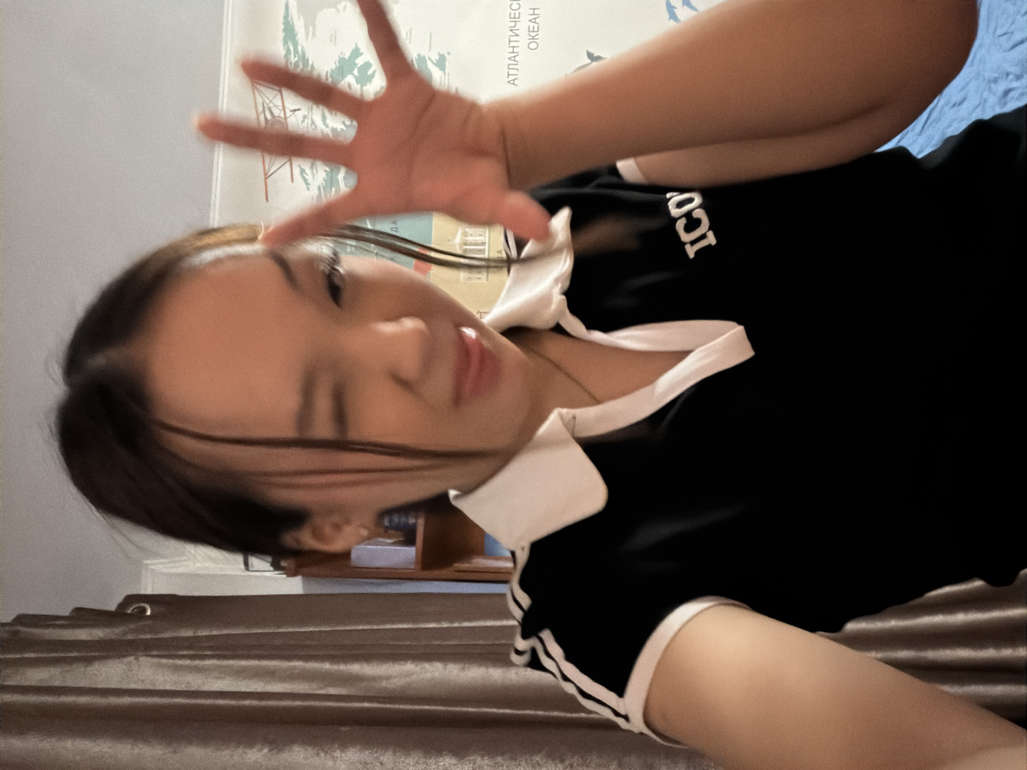 This image captures a young East Asian female in an indoor setting within the city of Unknown, Russia. The subject is depicted from the chest up, making a playful expression by winking with her right eye and sticking out her tongue. Her left hand is raised with fingers splayed, partially obscuring her face and exhibiting motion blur. She wears a black short-sleeved polo shirt with a white collar, white trim on the sleeves, and the white letters "ICO" on the chest. A small earring is visible in her left ear and she wears a thin necklace. The background on the left features a textured brown curtain. On the right, a wall-mounted map is visible, displaying landmasses, an illustration of an old biplane, and Russian text including "АТЛАНТИЧЕСКИЙ ОКЕАН" (Atlantic Ocean). A wooden bookshelf is partially visible behind her left shoulder, and a blue fabric element is present at the bottom right. The ceiling is white.