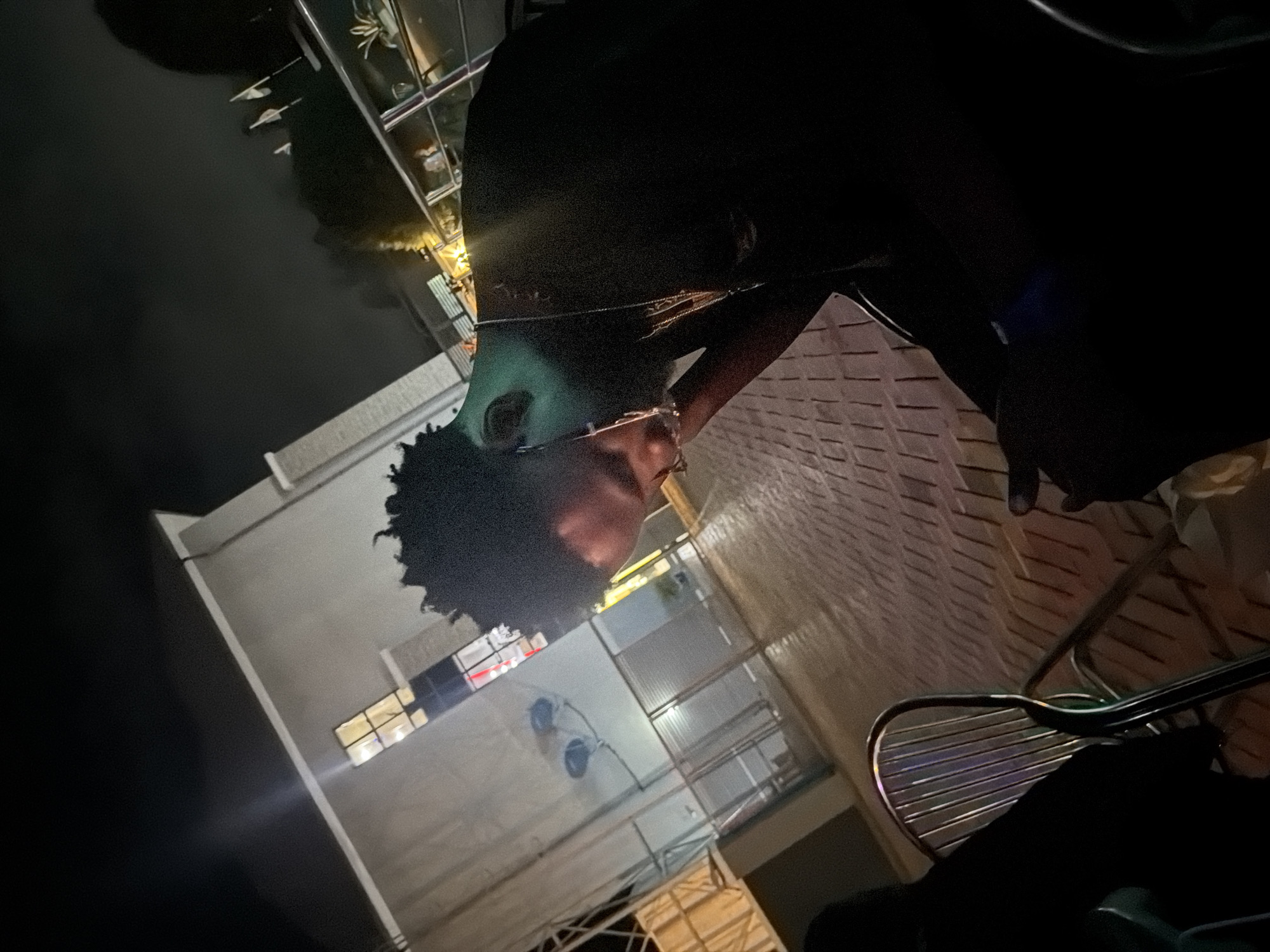 An outdoor scene at night, likely in Bukuru, Nigeria, captures a young Black man seated at a table. He is leaning forward, head bowed, and appears to be wearing glasses. His skin is illuminated by a greenish light, possibly from nearby lighting or a filter. The background features a building with windows, a paved ground, and some chairs. The overall lighting is dim, indicating it is nighttime.