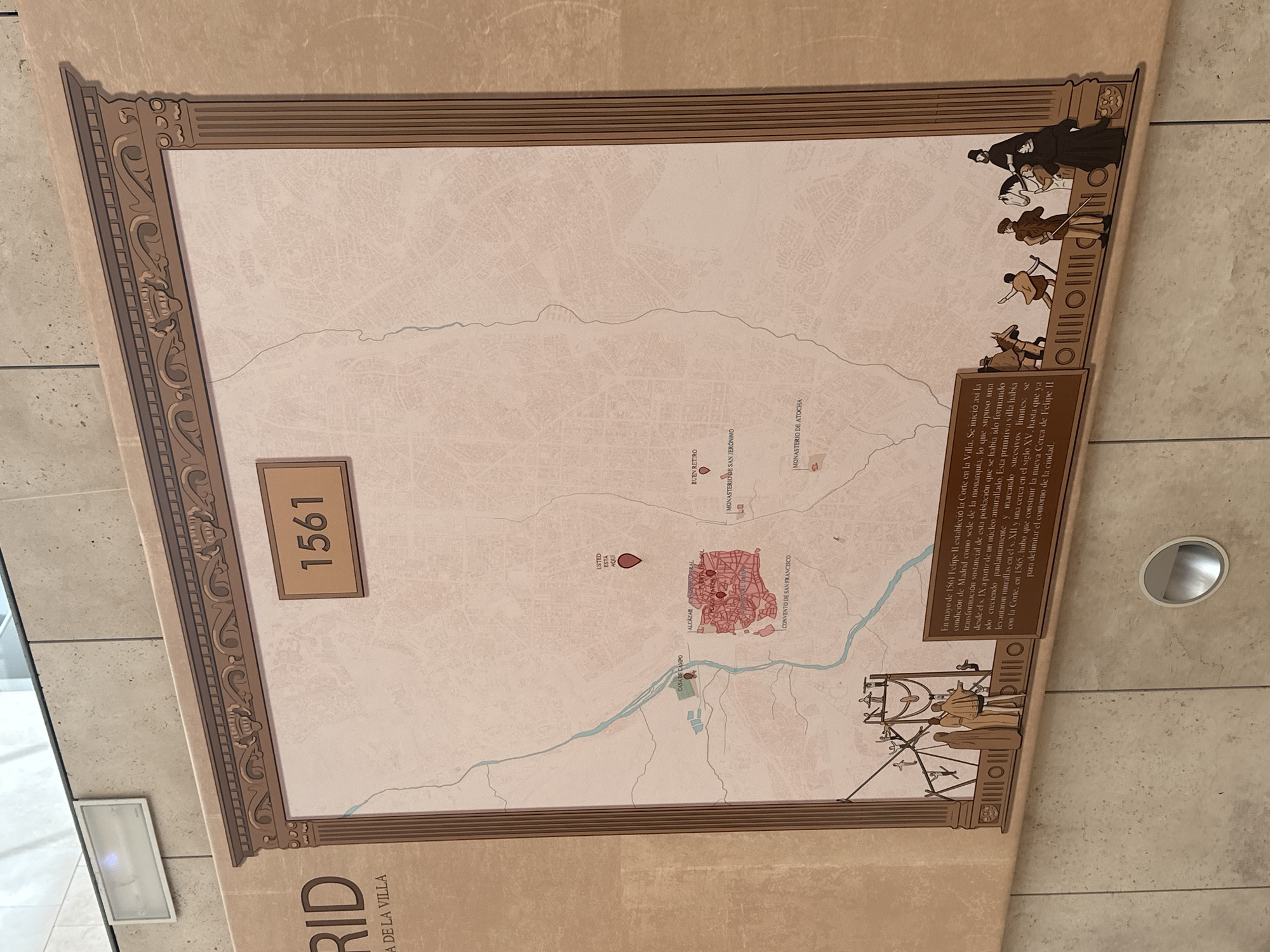 The image shows a framed map of Madrid, Spain, dated 1561. The map depicts the city's layout with key locations marked, including Alcázar Real, Plaza Mayor, and Convento de San Francisco. Several historical figures in period attire are illustrated along the bottom edge of the map, some on horseback, and others walking. A text box below the map provides historical context about Madrid's development in 1561. The overall presentation suggests an informational display in a museum or public space.