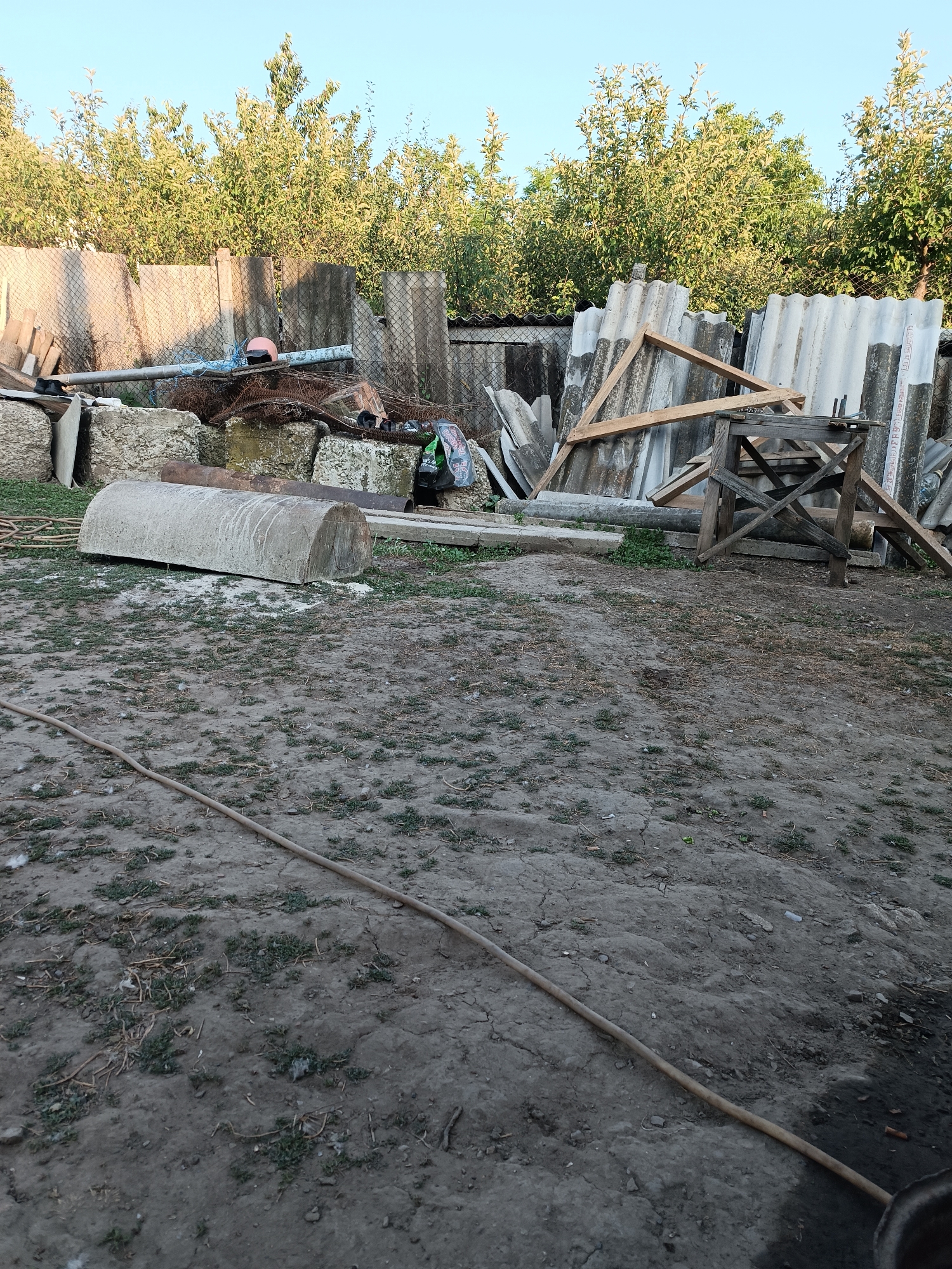 The image depicts a cluttered outdoor scene, likely a yard or storage area, in Leușeni, Moldova. A large, half-cylindrical concrete object lies on the ground in the foreground, partially covered by a thin layer of dirt and sparse vegetation. A long, flexible hose snakes across the ground, extending from the lower left towards the middle right of the frame.
In the background, a pile of assorted materials is haphazardly stacked. This includes various sheets of corrugated roofing material, some leaning against a fence, others supported by wooden beams. A wooden sawhorse-like structure stands among the debris. Interspersed with the building materials are pieces of concrete, metal pipes, and what appears to be tangled wire or mesh. A bright pink helmet rests on a pipe.
Behind the accumulated items, a dense line of green trees forms a backdrop, with a clear, pale blue sky visible above. The lighting suggests it is either early morning or late afternoon, with the sun casting soft shadows. The overall impression is one of disuse and accumulation of discarded items, possibly indicating a renovation project or a junkyard. No people are visible in the image. There is no discernible text in the image.
