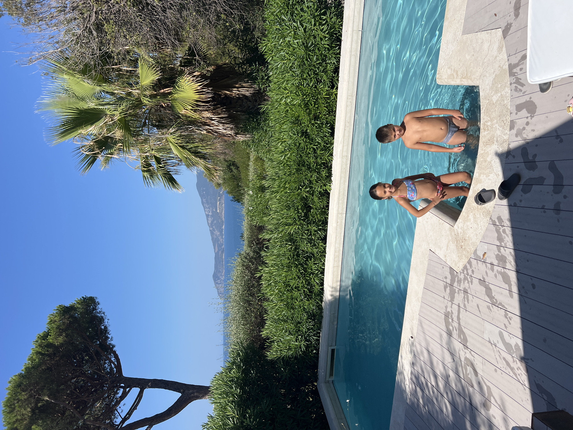 In Orbetello, Italy, two children stand in a swimming pool by a wooden deck. The girl, on the left, wears a floral bikini, and the boy, on the right, wears gray swimming trunks. They are positioned in the shallow end of the pool. Several pairs of flip-flops are visible on the deck. The pool is surrounded by lush green foliage and a palm tree. In the background, the blue sea and distant hills are visible under a clear blue sky. The scene captures a sunny day at a poolside location.