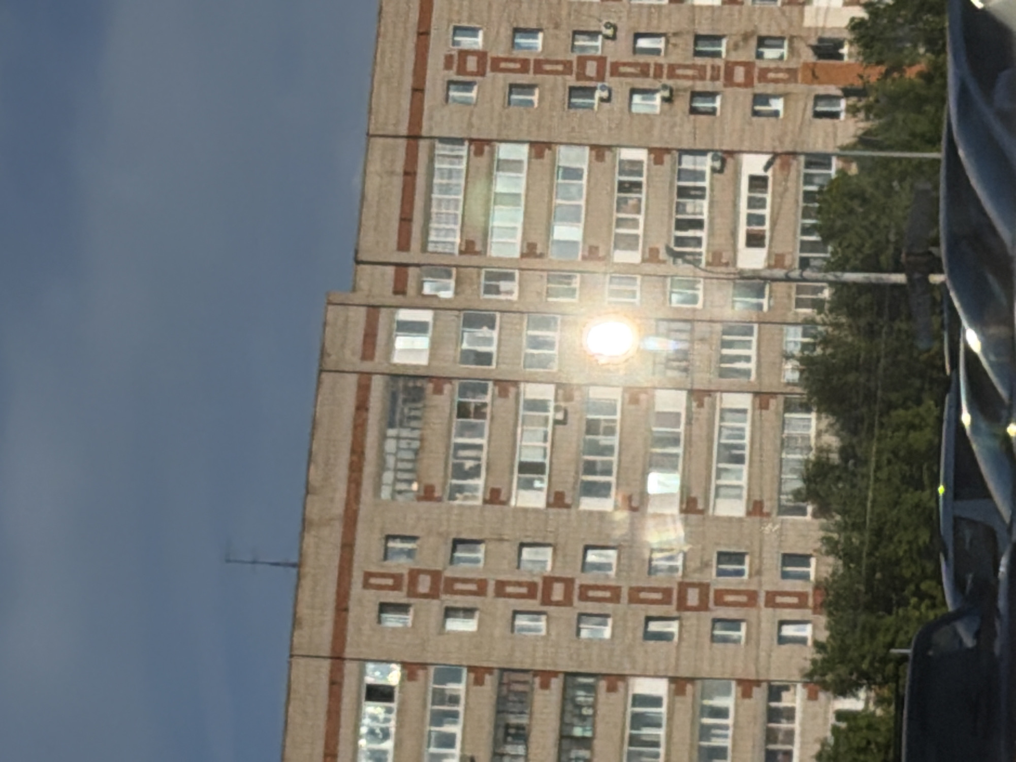 The image, viewed in its correct upright orientation, captures a daytime scene dominated by a large, multi-story residential building. This building, a prominent subject on the left side of the frame, displays a light-colored facade, likely concrete or panel, accented with distinct horizontal and vertical brown rectangular decorative patterns between its numerous windows. Many windows feature white frames and reflect the bright sunlight, with some sections appearing as continuous vertical glass strips or enclosed balconies.
A strong, circular sun glare is prominently visible on the building's facade, indicating clear and sunny weather. Above the building, a slender antenna or mast extends into a vast expanse of clear, light blue sky that occupies the right portion of the image. Green foliage from trees is visible in the lower right, partially obscuring the building's lower floors.
In the immediate foreground on the far right, parts of a dark-colored vehicle, likely a car, are visible, its reflective surfaces also catching the sunlight. A dark pole or vertical structure is partially discernible in this area. No people or visible text are present in the image. The building's architectural style is characteristic of urban residential structures found in cities like Izhevsk, Russia.