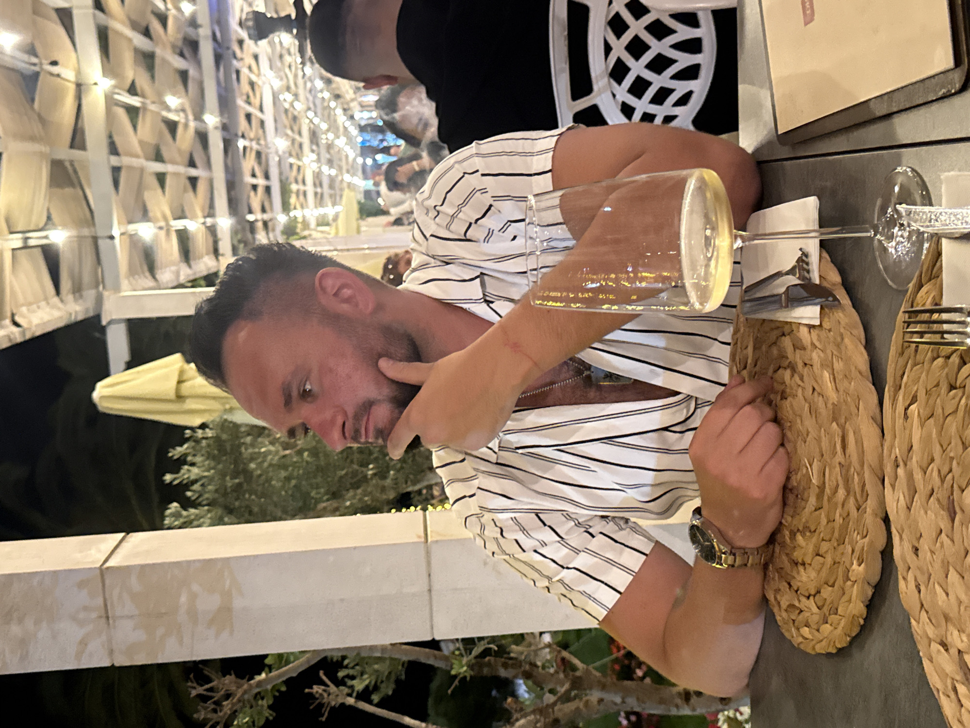 The image depicts an outdoor restaurant scene in Rafailovići, Montenegro, during the evening. A male individual, appearing to be in his 30s or 40s, is seated at a dark-colored table, positioned slightly off-center to the left. He has dark, short-cropped hair with a fade and a short beard. He is wearing a short-sleeved, open-collared shirt with vertical black and white stripes. A metallic chain necklace is visible at his open collar. A watch with a metallic band and a dark face is on his left wrist. A faint reddish mark is present on the inside of his right forearm. He is looking towards the left side of the frame, with his right hand resting on his chin in a pensive gesture. His left forearm is resting on a round, woven placemat made of a natural material, possibly straw or wicker.
On the table in front of him, to his right, is a clear wine glass containing a pale yellow liquid, consistent with white wine, showing reflections of ambient lights. Cutlery (a fork and knife) wrapped in a white napkin is partially visible to the left of the wine glass. A brown menu or booklet is situated on the right side of the table.
The background reveals an outdoor dining area under a pergola or canopy adorned with numerous small string lights, creating a soft illumination. Pillars, likely part of the restaurant's structure, are visible to the left of the man. Foliage and bare branches of trees are present in the left background, and an unlit yellow patio umbrella is seen further back. Other patrons are discernible as blurry figures in the background, suggesting a populated dining environment. Another individual in a dark shirt is seated with their back to the camera in the right mid-ground.