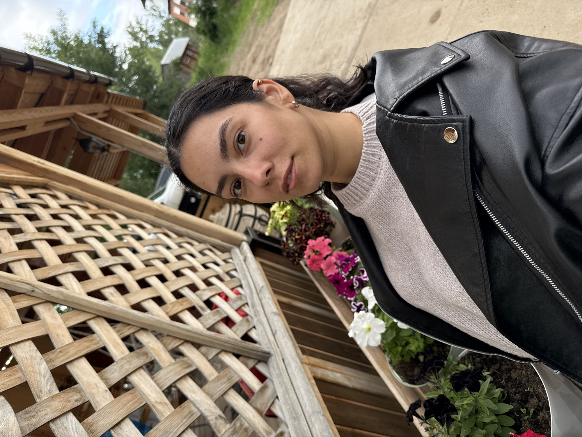 An outdoor portrait depicts a woman in her 20s or 30s, positioned under a wooden structure, possibly a pergola or porch. She has dark hair, styled to appear somewhat wet or sleek, and brown eyes, looking directly at the camera with a neutral to slight smile expression. She wears a light-colored, possibly off-white or light grey, knitted sweater with a crew neck, layered under an open black faux leather or leather jacket featuring a wide collar with a gold-toned snap button on the left and a silver-toned snap button on the right, and visible zippers. Small silver-toned hoop earrings are in her earlobes, and a silver-toned stud or ring is in the helix of her left ear. A small mole is visible on her right cheek.
To the left of the woman, a prominent wooden lattice screen, constructed from interwoven light-colored wooden strips, forms a wall or partition. Below this lattice, a horizontal wooden beam and vertical wooden slats create a lower wall. Beneath these wooden structures, a planter box holds various flowering plants, including white, pink, and purple petunias, alongside darker leafy plants with visible soil.
The background reveals more elements of the wooden structure overhead, including beams, supports, and a metal gutter system. A dark-colored lantern-style light fixture hangs from the wooden ceiling. Beyond the immediate structure, the scene transitions to an outdoor environment with blurred green foliage (trees and bushes) against an overcast or bright sky. A paved or dirt path extends into the distance, leading to several small, simple buildings or houses. A white vehicle is partially visible and blurred behind the wooden structure on the far left. The specific geographical location in the city of Chaikovsky, Russia, cannot be confirmed from the visual information present in the image.