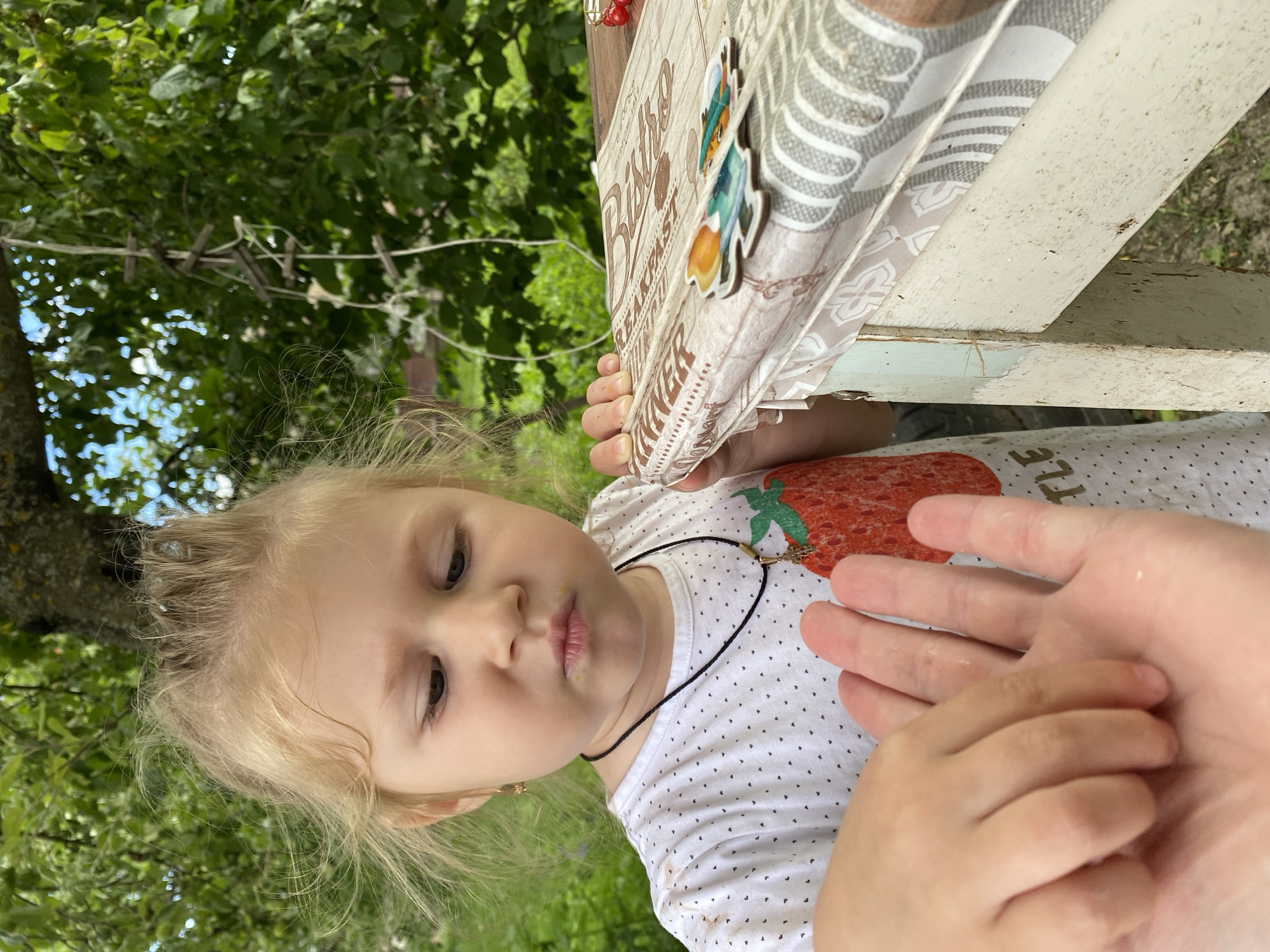 This is a medium shot, outdoor photograph taken in a garden setting, likely in Маринопіль, Ukraine. The image is oriented vertically, with the subjects appearing upside down.
The primary subject is a young blonde-haired girl with dark eyes, looking downwards with a pensive or curious expression. She is wearing a white t-shirt with a repeating small black heart pattern and a large strawberry graphic. Around her neck, she wears a thin black cord with a small cross pendant. Her left ear is adorned with a small golden earring. Her left hand is reaching towards an adult's open right hand, which is in the foreground. The adult's hand is pale and appears to be offering something or inviting interaction.
In the background, there are lush green trees and foliage, suggesting a warm, sunny day. Part of a wooden structure, possibly a table or bench, is visible to the right, adorned with a decorative panel. This panel has a light-colored textured surface with text and graphics. Visible text on the panel includes "BISTRO", "BREAKFAST", and "DINNER". There are also two small wooden cutouts attached to the panel, one depicting a fox-like character in a green hat and another with a brown animal head. A clothesline with several wooden clothespins is strung across the branches in the upper left portion of the frame.
The lighting suggests it is daytime, with natural sunlight filtering through the leaves. The overall mood is casual and domestic, capturing a tender moment between a child and an adult in an outdoor environment.