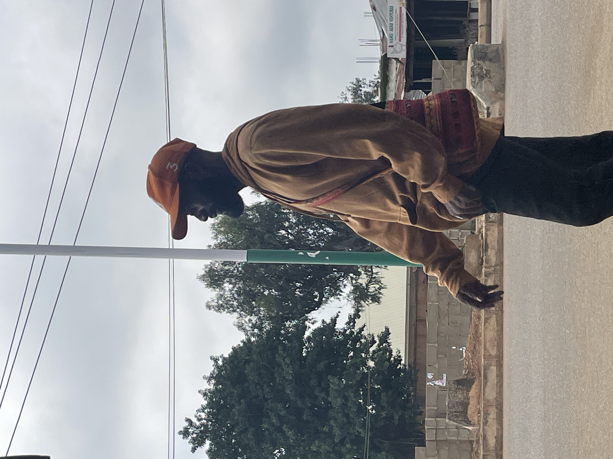 A man walks along a paved road in Bukuru, Nigeria. He is wearing an orange baseball cap with the number "3" on the side, a tan hooded sweatshirt, and dark pants. A colorful woven bag is slung across his chest. He has a beard and is looking to his left. In the background, there are trees, utility poles, power lines, and buildings, including one with a sign that reads "NAMU AGRO VENTURES." The sky is overcast.