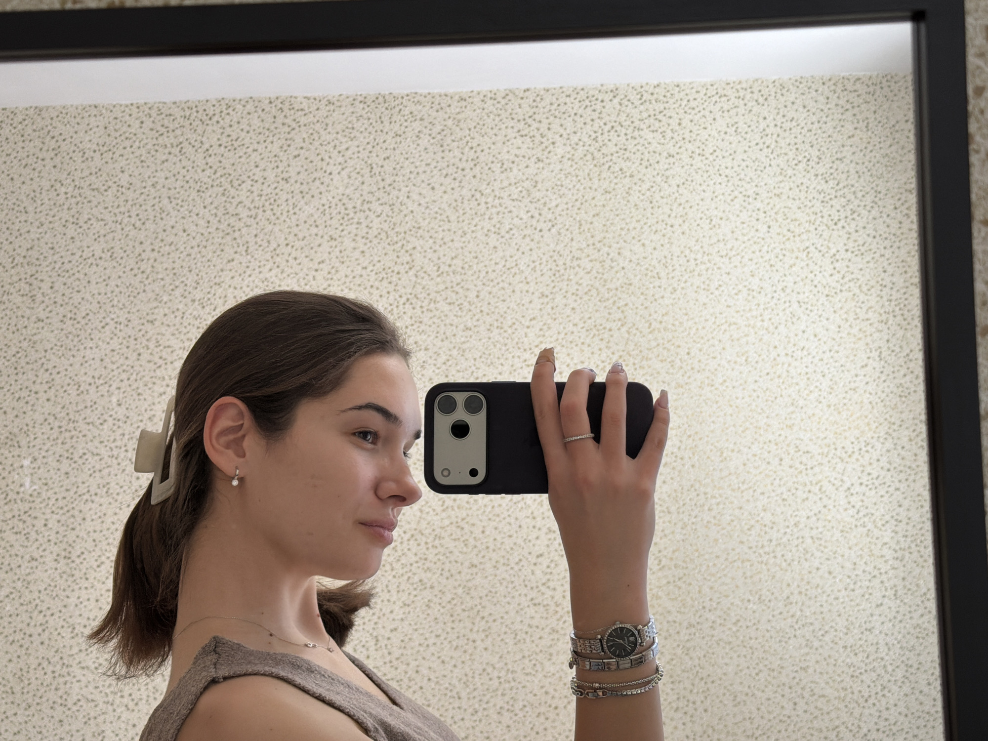 A young woman is taking a selfie in front of a mirror. The mirror has a black frame and reflects a wall with a speckled pattern. The woman is holding a black smartphone in her right hand, obscuring part of her face. She has brown hair pulled back with a beige claw clip and is wearing a small earring, a delicate necklace, a ring on her finger, and a watch with multiple bracelets on her wrist. The location is possibly in Sofia, Bulgaria, but there is no visual evidence to confirm this.