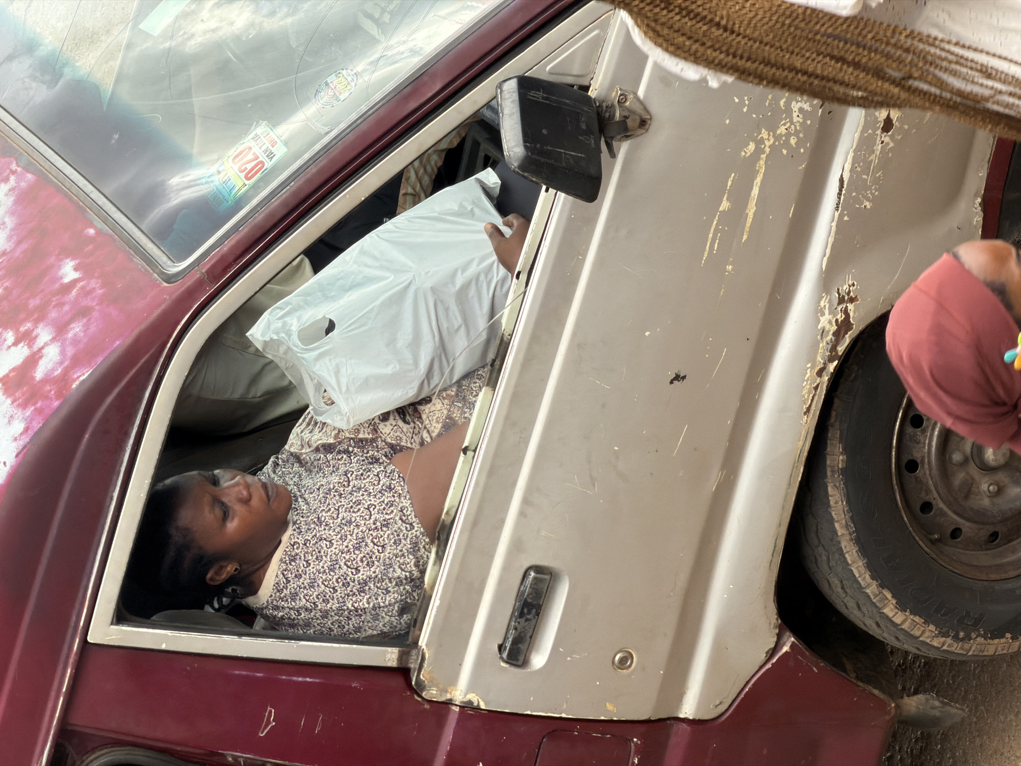In Bukuru, Nigeria, a woman is seated in the driver's seat of a weathered maroon and silver van. She is looking down, holding a large white plastic bag in her lap. Her hair is styled in braids and she is wearing a patterned dress. The van's exterior shows signs of wear and tear, including rust and scratches. Through the windshield, stickers are visible. The background is blurred, suggesting the photo was taken outdoors.