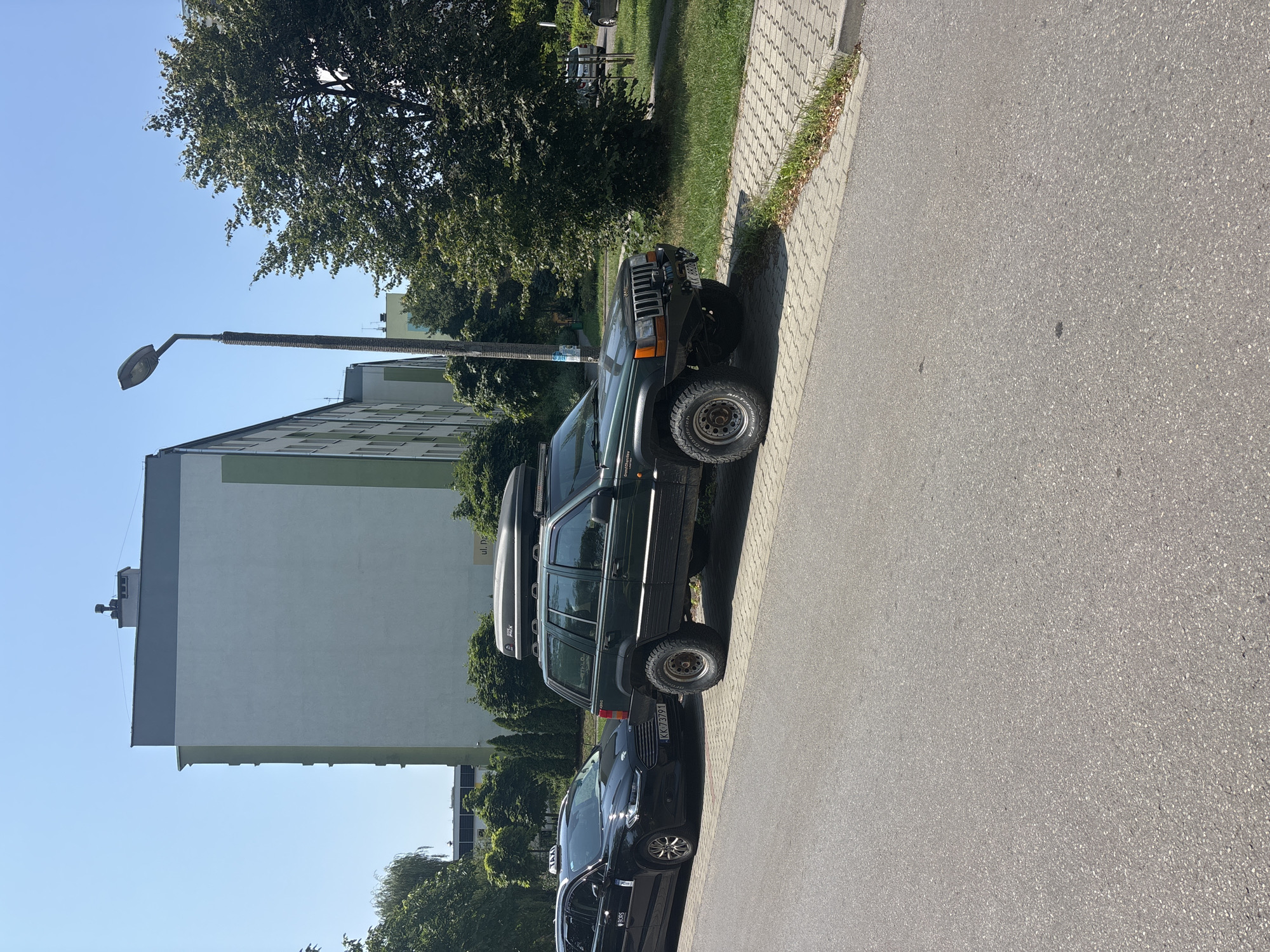 A dark green, lifted Jeep Grand Cherokee is parked on the side of a residential street in Krakow, Poland, next to a black taxi. The Jeep has a roof-mounted cargo box and off-road tires. The taxi has a "TAXI" sign on its roof and a license plate that reads "KK 73791". In the background, there are tall, light-colored apartment buildings and lush green trees. The sky is clear and blue, indicating a sunny day. The street is paved with asphalt, and there is a sidewalk made of interlocking pavers. The area appears to be a typical urban environment with residential buildings and street parking.