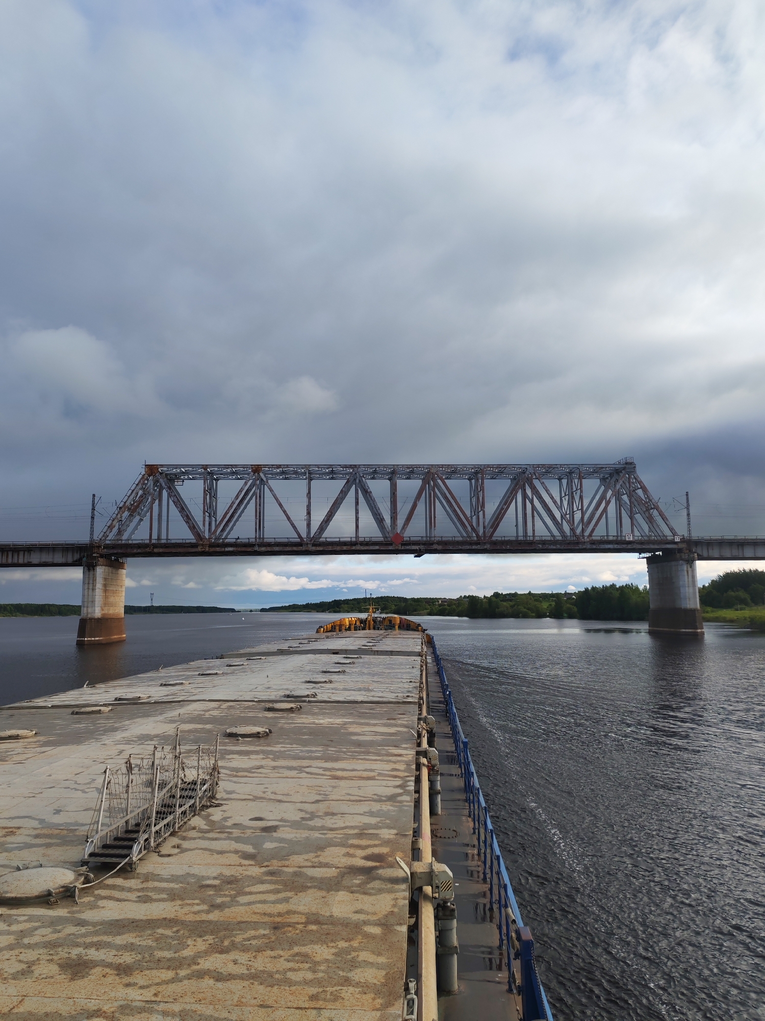 The image depicts a view from a barge or large boat, possibly in the vicinity of Tarkanovo, Russia, based on the presence of the bridge. The barge has a rusty, weathered surface with hatches and metal railings. A truss bridge with train tracks spans the river, supported by concrete pillars. The river surface shows ripples and wake, suggesting movement. Trees and vegetation are visible on the riverbanks. Overcast sky conditions prevail. No people are visible.
