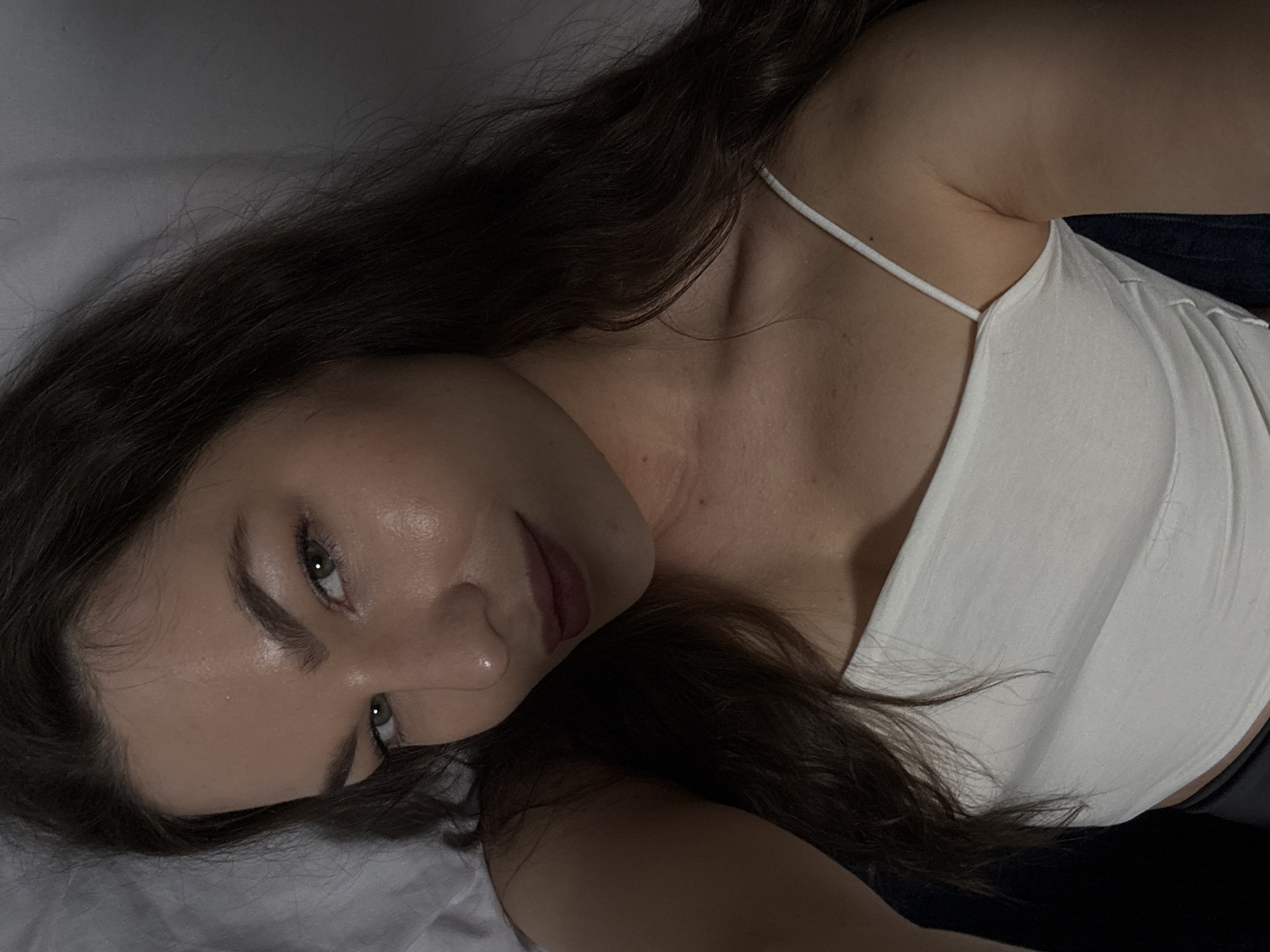 The image is a close-up selfie of a young woman with long, dark brown hair and green eyes. She is wearing a white spaghetti strap top. Her skin appears dewy and lightly made up. The lighting is dim, casting shadows on her face and hair. The background is blurred and appears to be a bed with white sheets, suggesting an indoor setting. There is no discernible location in the city of Chaikovsky, Russia.