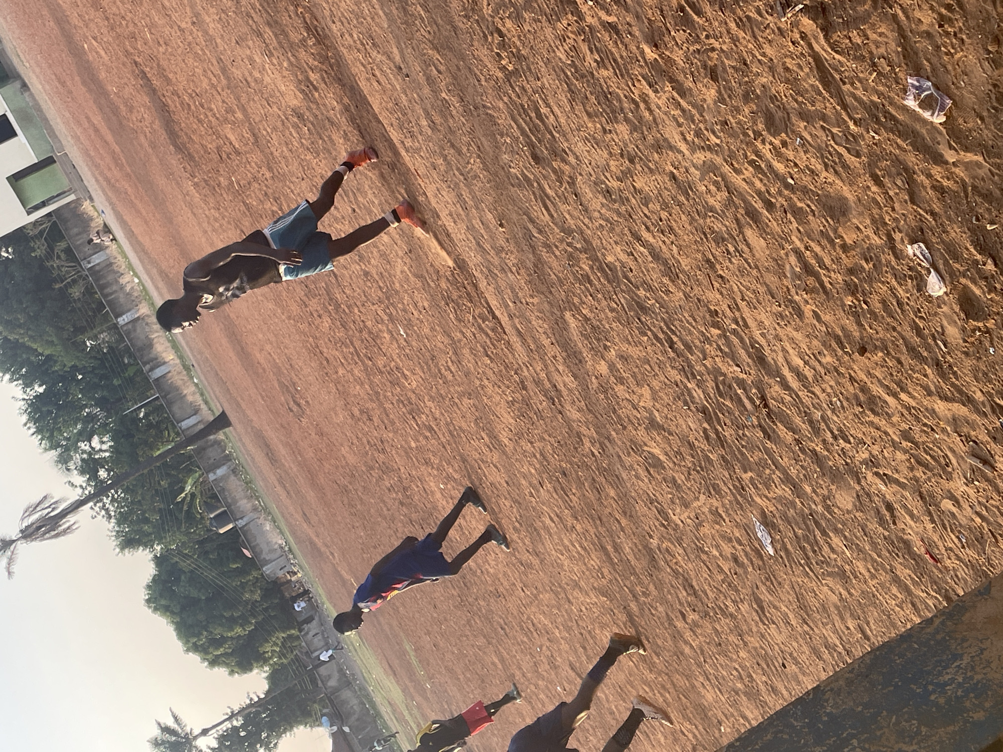 The scene shows an outdoor space that appears to be a dry, dirt or clay-like field. There are several young men, likely athletes or runners, present. One man, in the upper left, wears a dark top, light blue shorts, and bright orange running shoes. Another man wears a blue and red shirt, and dark shorts. Two additional men are visible in the lower portion of the frame, one wearing red shorts and another wearing blue shorts. The background includes a building and trees, suggesting this location is in a developed area, most likely in Akwanga, Nigeria. There is also debris scattered on the field. The image appears to have been taken in the late afternoon or early morning due to the angle of the sunlight.
