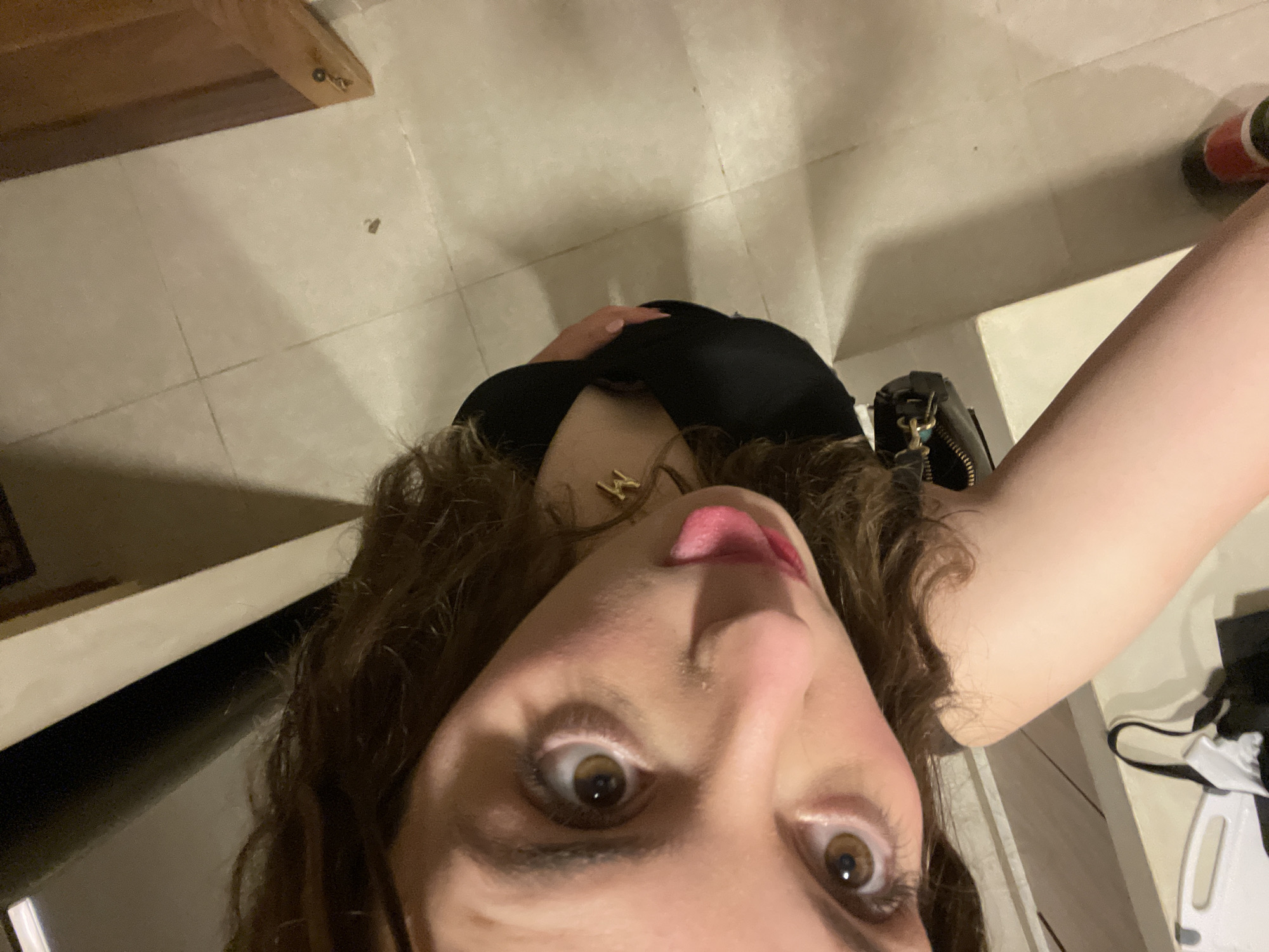 A top-down selfie, taken from a low angle, features a woman indoors. She has brown curly hair, fair skin, and brown eyes. Her tongue is sticking out slightly. She is wearing a black top and a gold necklace.
The background includes light-colored tiled flooring and parts of furniture, including a wooden table leg and a white storage unit. A black purse and what appears to be a red and black bottle are also visible on the floor near the storage unit. The image offers no specific information to confirm the location in Brisas de Zicatela, Mexico.