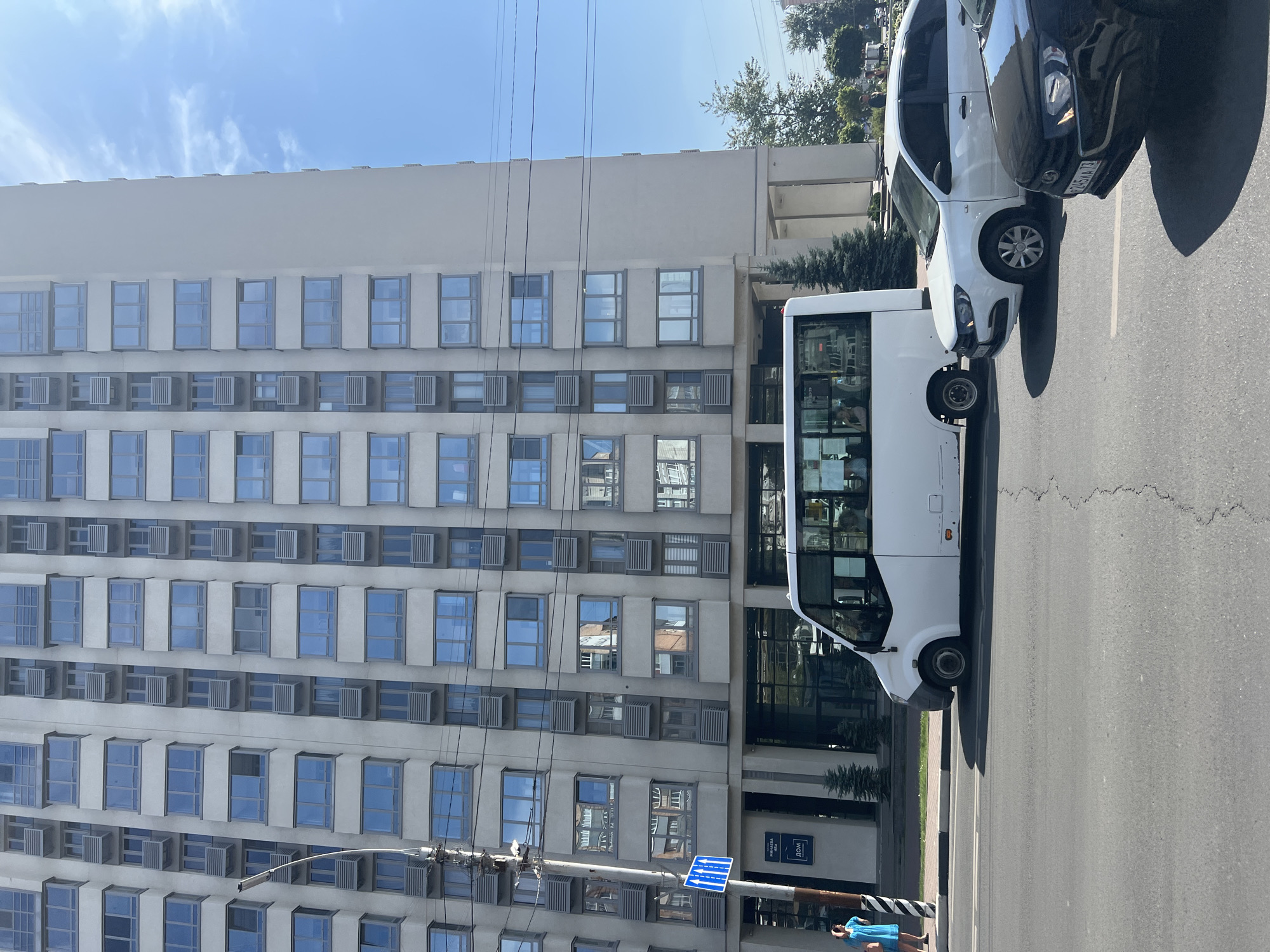 An outdoor, eye-level, medium shot captures a street scene in front of a modern, multi-story apartment building. The building is light gray with numerous windows, each featuring gray frames and what appear to be air conditioning units beneath them. The sky above is bright blue with a few wispy clouds. In the foreground, a paved road is visible with traffic lines. A white minibus is parked on the right side of the road, facing left. To the right of the minibus, a white sedan is also parked. Further to the right, the front of a black sedan is visible. On the left side of the image, a blue traffic sign with white arrows is mounted on a pole. A woman in a blue dress stands to the left of the traffic sign. The ground in front of the building is paved, and several small evergreen trees are planted along the sidewalk. The overall scene suggests a typical urban environment on a clear day.
