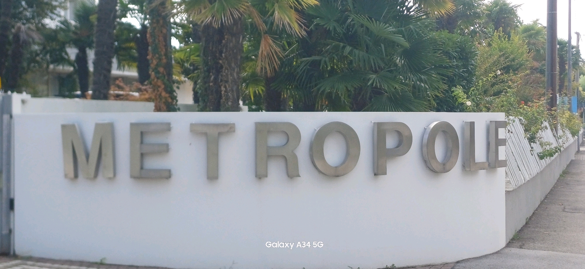The image shows a white wall with metal letters spelling "METROPOLE" in Abano Terme, Italy. Behind the wall is lush greenery including palm trees and bushes. The wall itself is a light grey concrete base with a white facade. A sidewalk runs along the right side of the image, adjacent to the wall and a decorative fence made of white vertical slats. The lighting suggests it is daytime with diffused light, possibly overcast. There are no people visible in the image. A watermarked "Galaxy A34 5G" is visible in the bottom right corner.