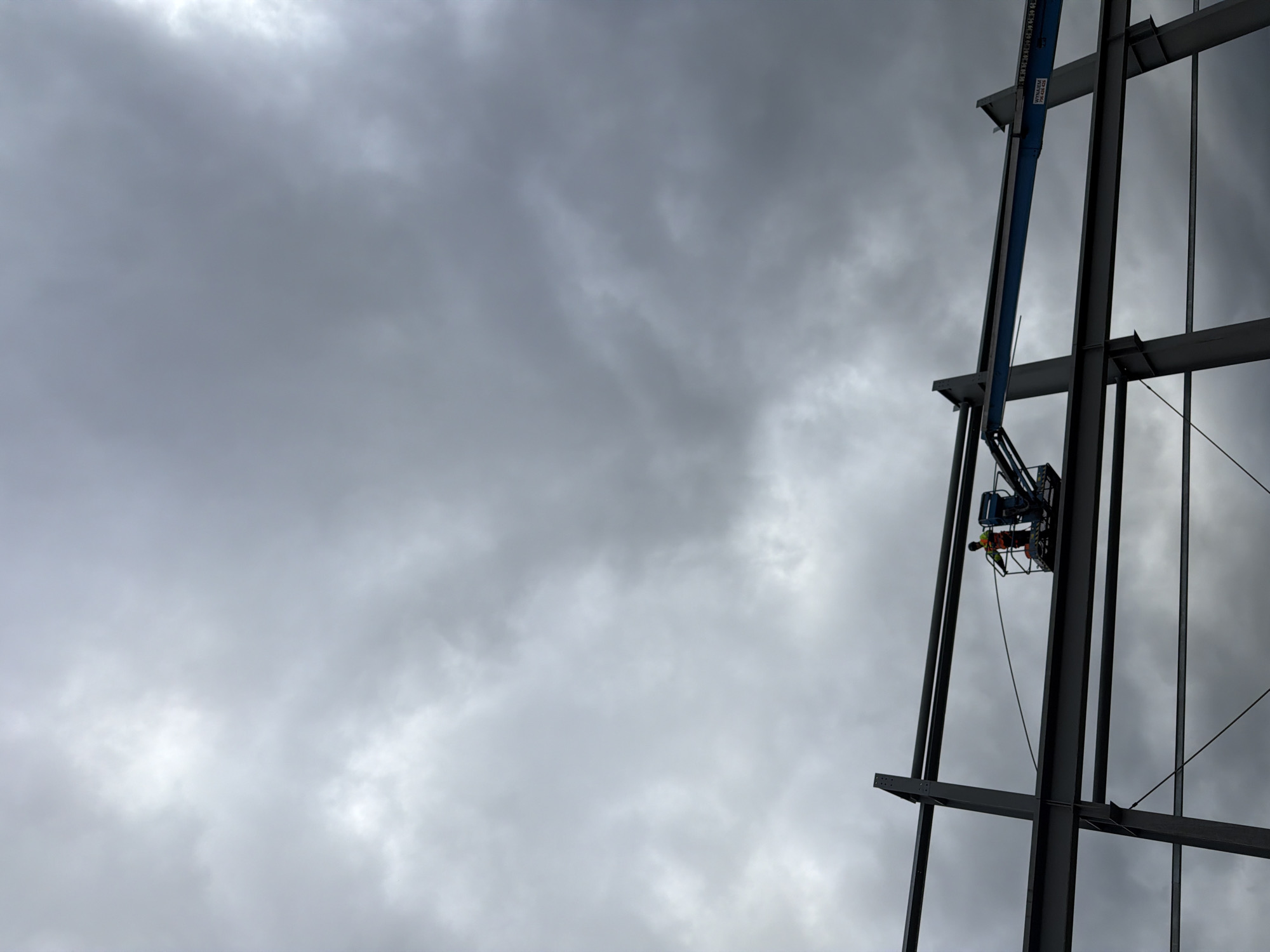 In the city of Stróża, Poland, a construction worker, wearing an orange safety vest, is operating a hydraulic boom lift at a construction site. The boom lift is positioned on the second level of a steel framework, which is partially erected. The worker is holding onto a wire that extends from the boom lift to the steel structure. The sky is overcast with dark clouds, suggesting an impending storm. The scene is a snapshot of an industrial construction project in progress.
