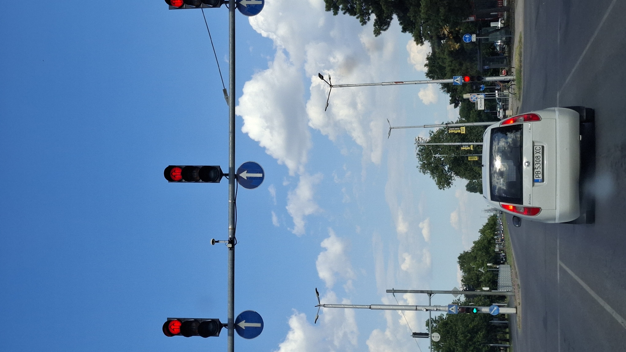 The image captures a view of a street in Plovdiv, Bulgaria, on a clear day with scattered clouds. The perspective is from a vehicle traveling on the road. In the foreground, the rear of a white Skoda Fabia is visible, with its license plate reading "PB 5308 XC". The car is emitting a faint white smoke from its exhaust.
Above the road, a series of traffic lights and signs are mounted on a metal bar. Three traffic lights are visible, all displaying red. Two blue circular signs with white upward-pointing arrows are positioned below the traffic lights. On the left side of the road, a traffic light is showing green.
Streetlights line the sides of the road, and several trees are visible, with dense foliage. In the background, other vehicles can be seen on the road, and a pedestrian crossing signal is present. The overall environment suggests an urban setting.
