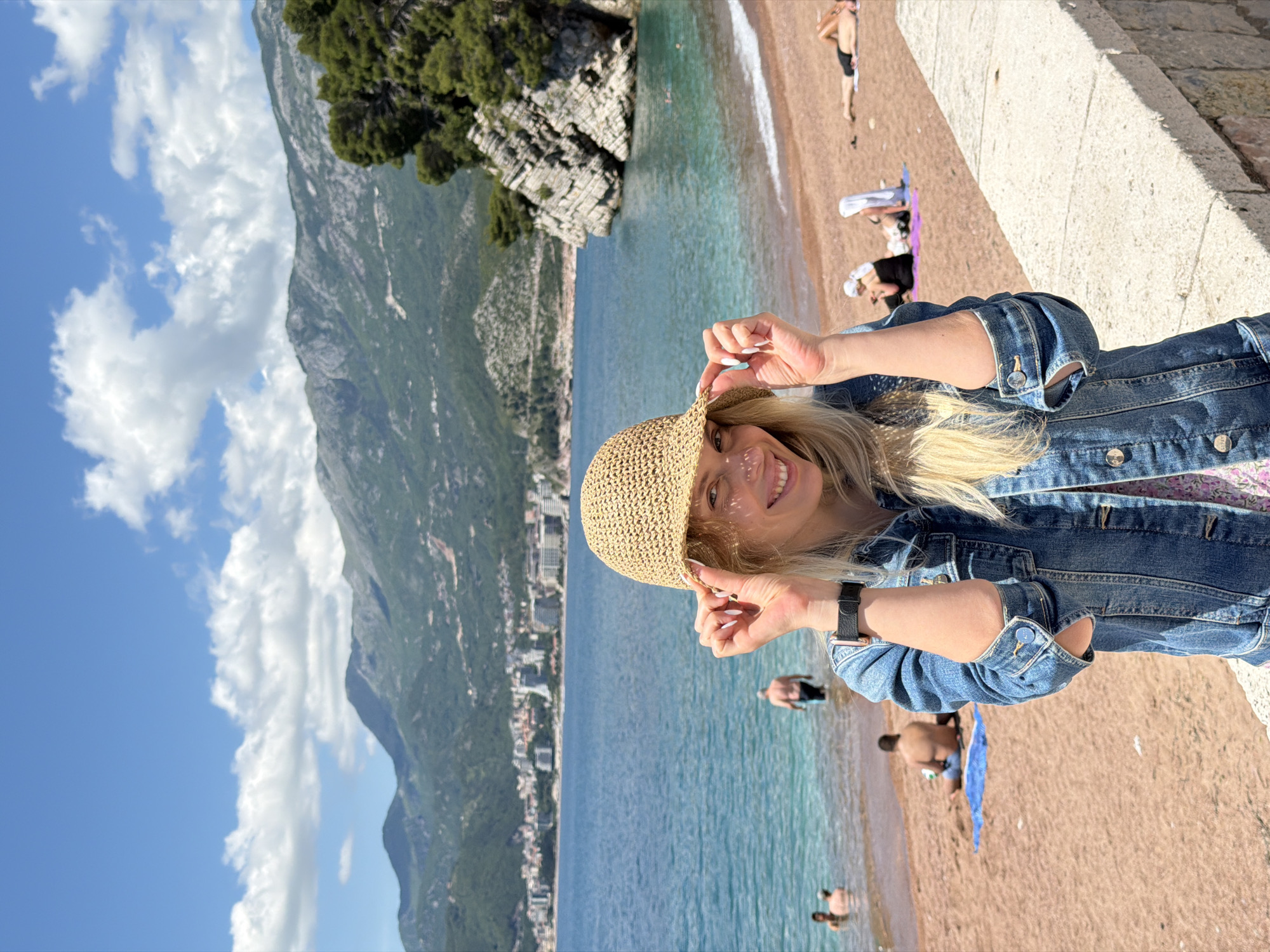 A woman is posing on a beach in Sveti Stefan, Montenegro, wearing a denim jacket and a straw hat. The background features the turquoise sea, a sandy beach with people sunbathing, and the city nestled against the mountains. The sky is blue with scattered clouds.
