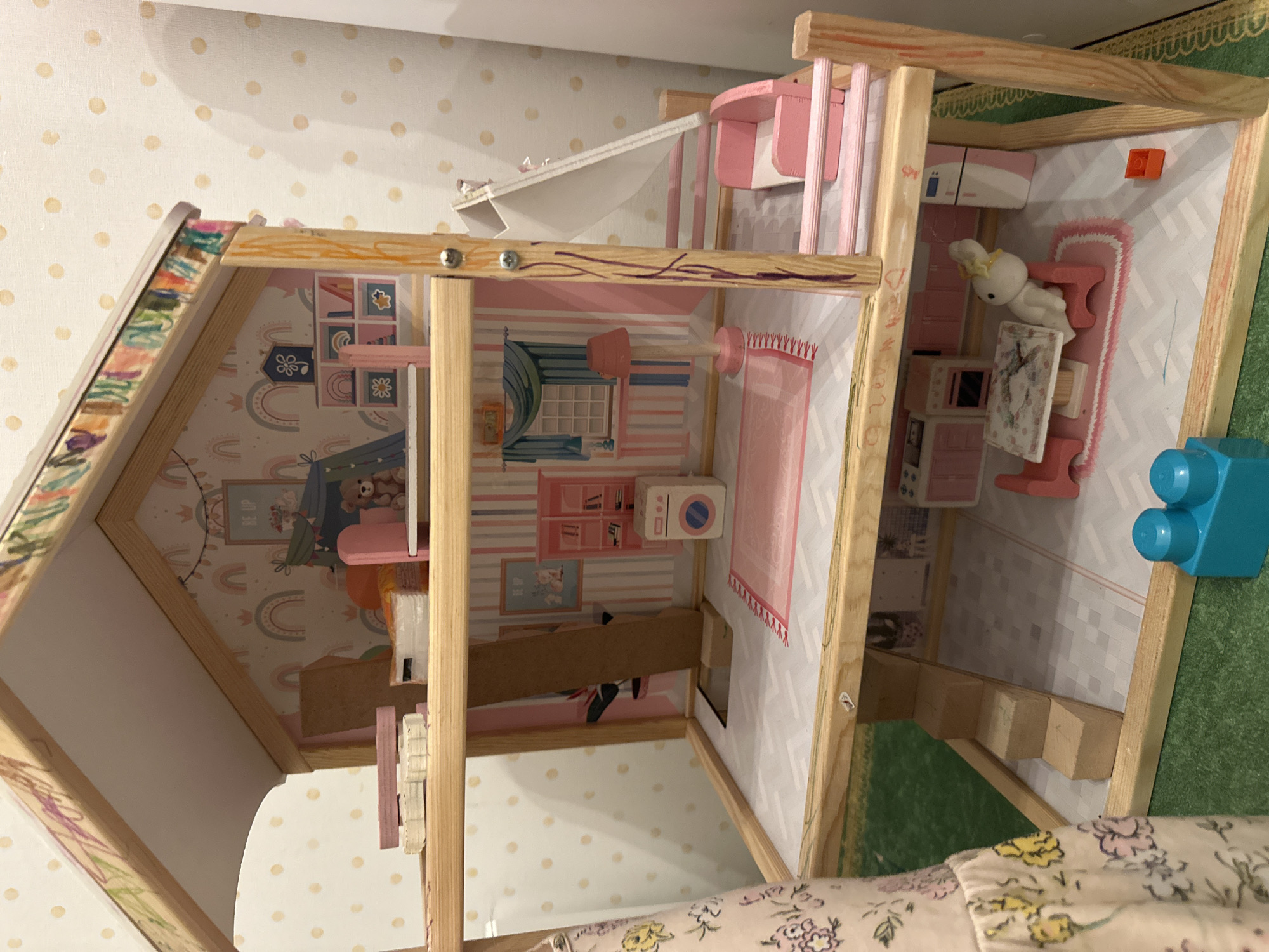 The image shows a dollhouse interior with three levels. The top level has a kitchen area with pink and white furniture and a small rabbit figurine. The middle level has a bedroom with a window and a pink-themed decor. The bottom level appears to be a child's room with a tent and rainbow-themed wallpaper. The dollhouse is made of wood with some pink accents. The location of the dollhouse is unknown but it is irrelevant to its description.