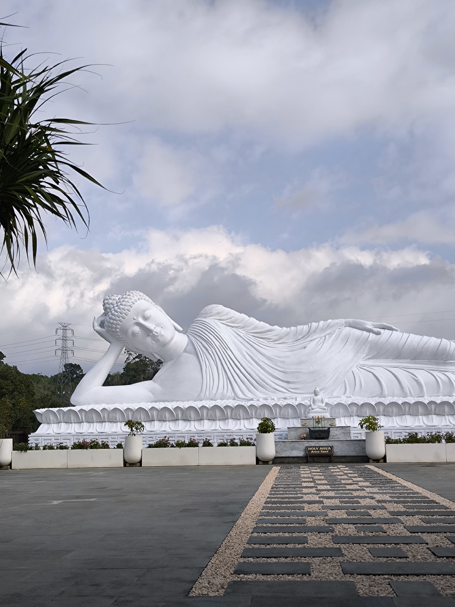 The image depicts a large white statue of a reclining Buddha, a significant landmark in Pupuan, Indonesia. The statue is positioned on a raised platform adorned with a decorative lotus petal motif at its base. In front of the statue, a paved pathway made of dark rectangular stones interspersed with small white pebbles leads towards a sign that reads "HOLY AREA Area Suci." The pathway is bordered by planters containing small green plants. The statue is set against a backdrop of a cloudy sky, with hints of blue peeking through. To the left of the statue, lush green foliage and a tall utility pole with power lines are visible, suggesting the statue is located in a somewhat natural or semi-rural setting within Pupuan. The overall scene conveys a sense of tranquility and reverence.