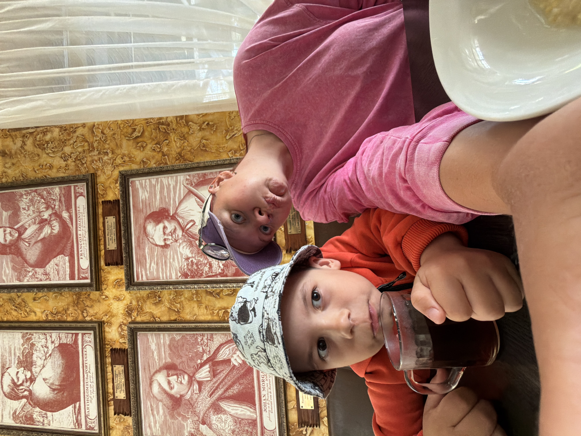 Two children are at a table inside what appears to be a cafe or restaurant. The child in the foreground wears an orange sweatshirt and a hat with dinosaur patterns and is drinking from a glass mug. Behind this child is another child wearing a pink sweatshirt and a purple baseball cap. The background features patterned yellow wallpaper adorned with framed prints of historical figures. A window with sheer white curtains is visible at the top of the image. The text on the framed prints includes names like "FRANCOIS LOLONOIS" and "JOHAN MORGA," along with descriptions in another language. The location is not specified as Chaykovsky, Russia, in the image.