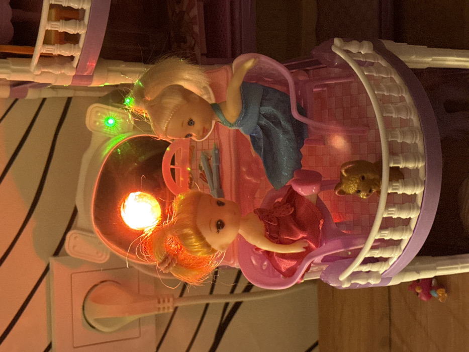 Two Barbie dolls are positioned inside a purple toy house with a light. One doll has blonde hair and wears a blue dress. The other doll has orange hair and wears a pink dress. A teddy bear figure is visible inside the house. The toy house is plugged into an electrical outlet. The walls behind the outlet have black stripes.
