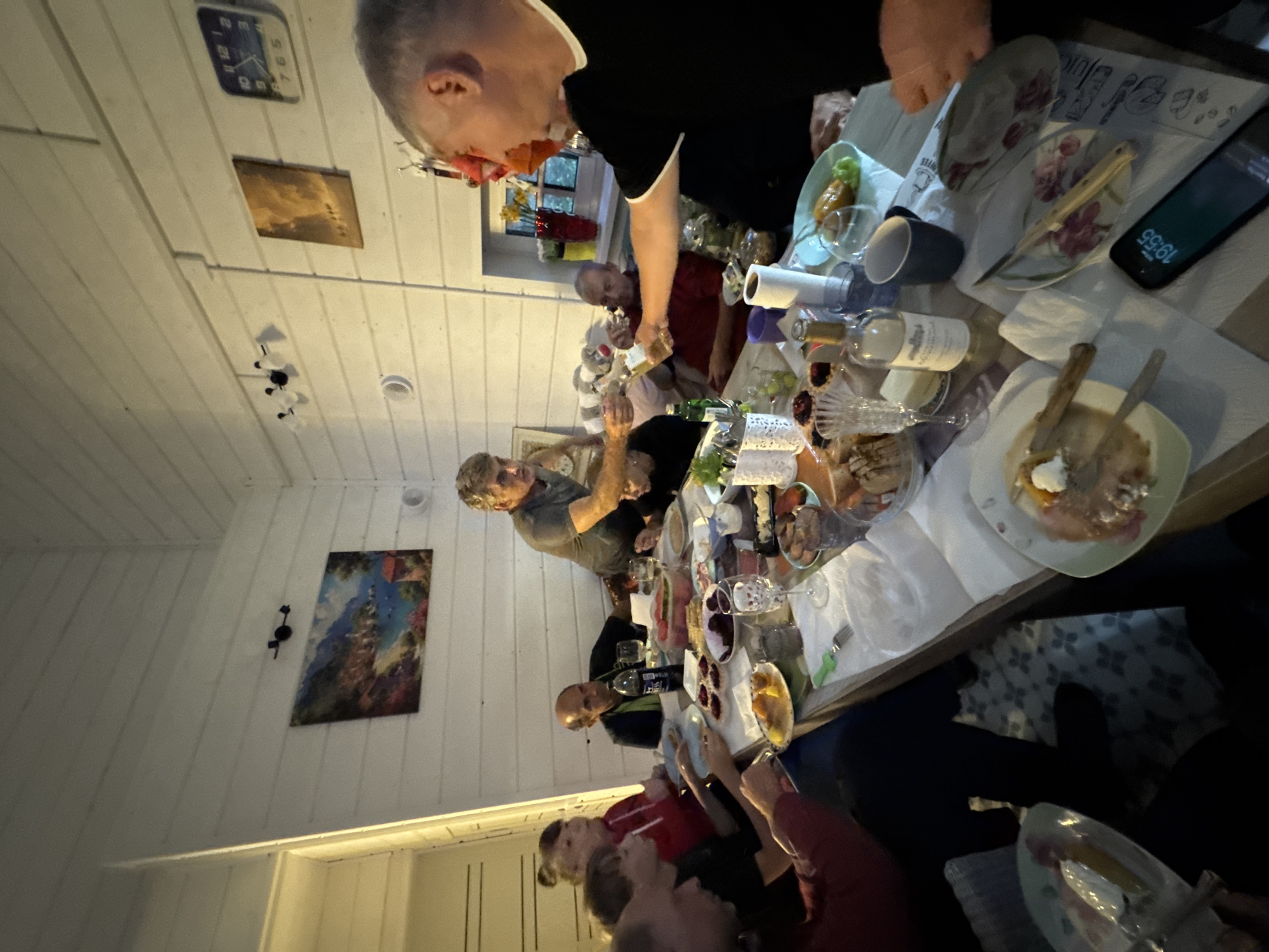 An indoor gathering is taking place around a long wooden table laden with food and drinks. Several people, appearing to be adults, are seated and standing, engaged in conversation and sharing a meal. The room has wooden paneled walls and a sloped ceiling. On the walls, there are framed artworks and a clock. The table is set with plates, glasses, bottles, and various dishes, including what appear to be pastries, fruits, and possibly a watermelon. One man in the foreground, wearing a black t-shirt, is in the process of pouring a drink from a bottle into a glass. Another man, in a gray t-shirt, is reaching towards the table. The overall atmosphere suggests a social event or celebration. The location is identified as Zelenaya Roshcha, Russia.