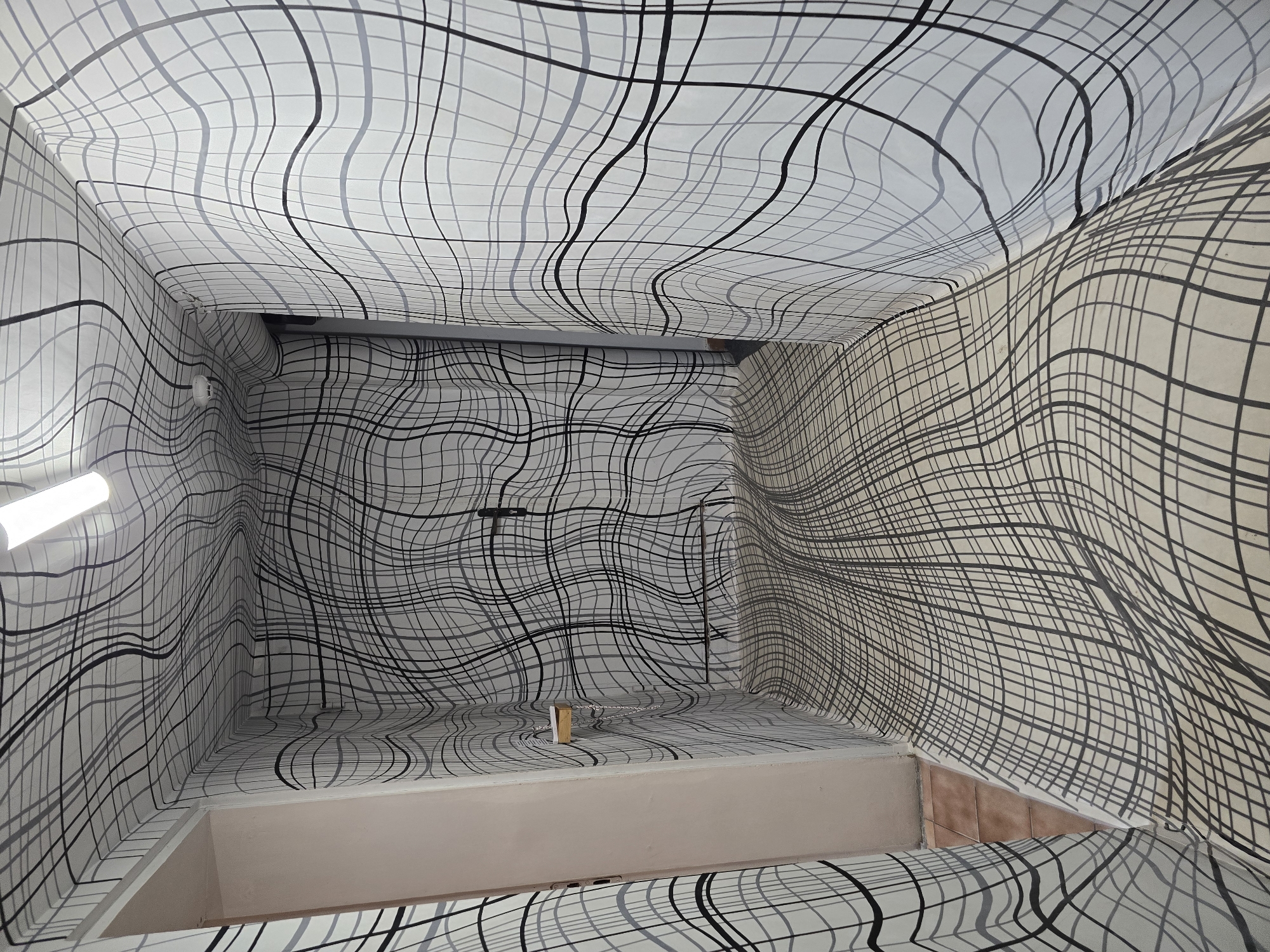 The image depicts a small, windowless room or space, possibly an elevator shaft or a storage area, completely covered in a white and black wavy line pattern wallpaper on all surfaces – the walls, ceiling, and floor. The lighting is provided by a single, bright fluorescent light fixture on one of the walls. In the center of the floor, there is a small, beige, rectangular block. Some metal components, such as a pipe or support beam, are visible along the ceiling and upper wall. There are no people or subjects visible in the image. The room appears to be in an unknown location.