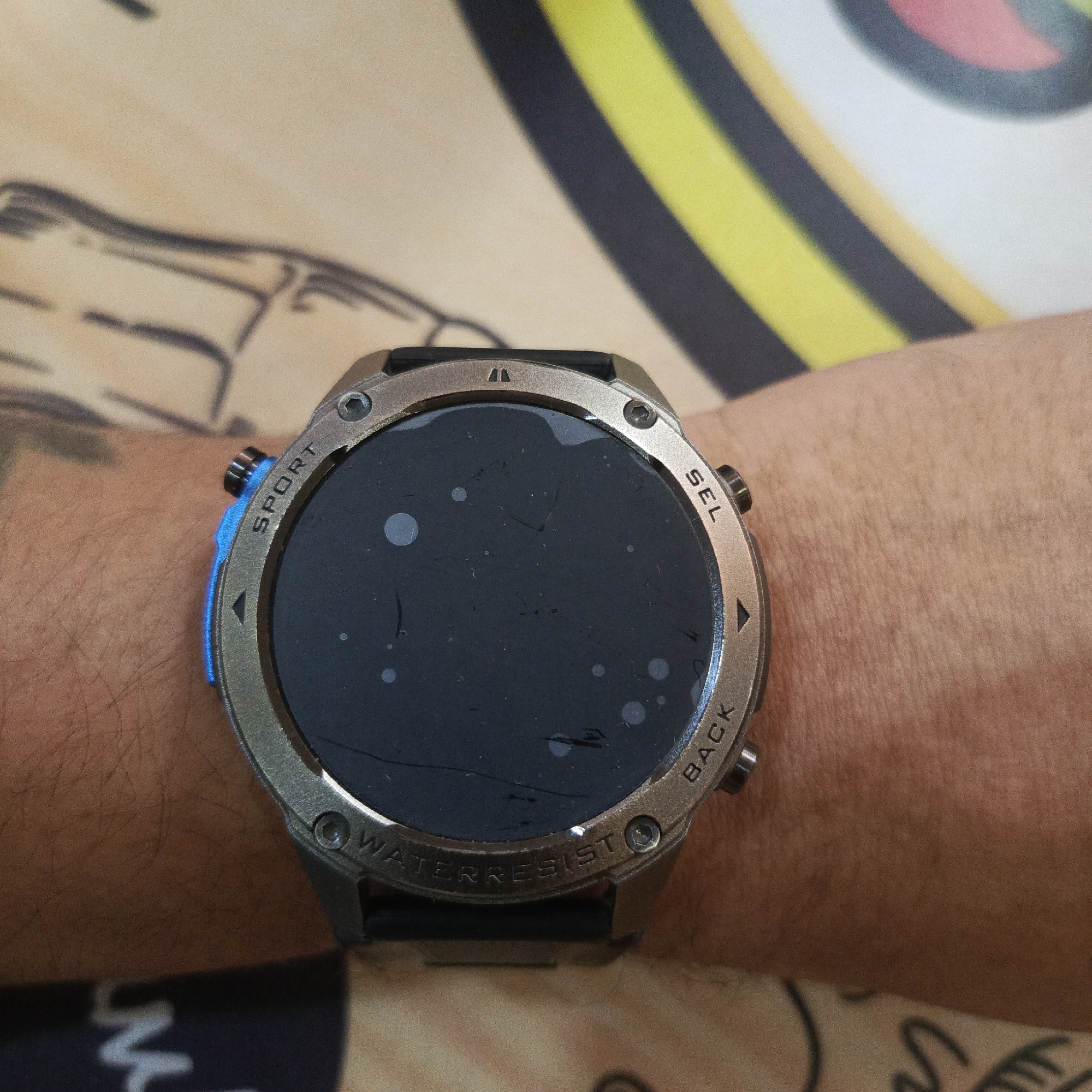 The image shows a close-up, eye-level shot of a smartwatch worn on a person's wrist. The watch has a metallic casing and a black screen with several light-colored circular smudges and some visible scratches. Text is embossed around the watch face, including "SPORT" on the left side, "SEL" on the top right, and "BACK" and "WATER RESIST" on the bottom. A blue illuminated button is visible on the left side of the watch. The person's wrist, covered in fine hairs, is visible below the watch. The background consists of a patterned fabric, featuring abstract, blurred shapes in yellow, black, and white. There are also faint dark lines visible on the fabric to the left of the watch. The scene is not identifiable as being in Los Teques, Venezuela.