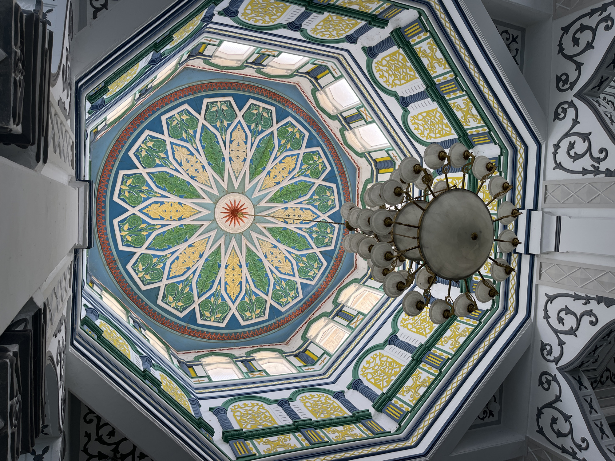 A low-angle shot captures the interior dome of a building, likely a mosque, in Keudekruenggeukueh, Indonesia. The octagonal dome features a circular, ornate design in the center, consisting of blue, green, and yellow floral patterns radiating from a central red star. The design is bordered by an orange band. Around the circular center are windows providing natural light. The surrounding structure is decorated with additional geometric and floral patterns in yellow, green, blue, and white. A large, multi-bulb chandelier hangs from the center of the dome. Gray ornate detailing is visible on the surrounding architectural elements.