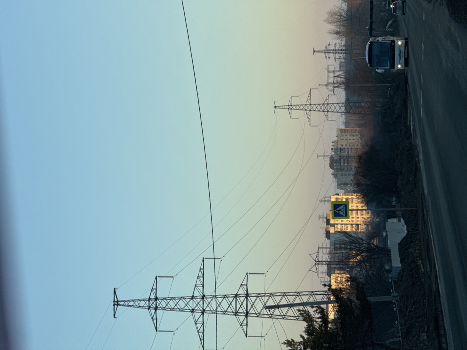 The image depicts an urban or semi-urban landscape in Chaikovsky, Russia, during what appears to be either early morning or late afternoon, characterized by low-angle sunlight. The sky transitions from light azure overhead to a pale yellow-orange at the horizon.
In the foreground, a two-lane asphalt road, possibly damp or reflective, stretches into the distance. A white tour bus with illuminated headlights is proceeding towards the viewer on the right lane, followed by a silver sedan, also with headlights on. To the left of the road, the ground consists of disturbed earth, appearing dark brown and uneven, with sparse, leafless deciduous bushes and trees. A dark green coniferous tree is partially visible on the far left edge.
Dominating the mid-ground and extending towards the background are numerous steel lattice electricity transmission towers (pylons), with multiple high-voltage power lines crisscrossing the sky. The largest tower is positioned prominently on the left. Behind these power structures and sparse vegetation, a cluster of multi-story residential buildings, likely apartment blocks, is visible. Several building facades, particularly on the left, are brightly illuminated by direct sunlight, appearing light yellow or beige. A blue square pedestrian crossing sign with a yellow border is affixed to a pole on the left side of the road. No individuals are directly visible.
