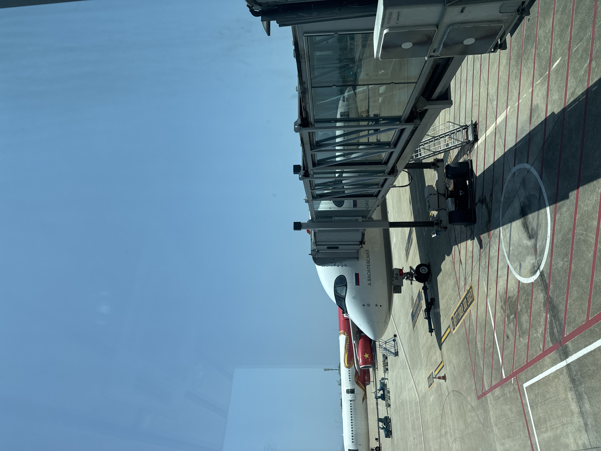 The image shows an airplane docked at an airport gate. The airplane is white with red and gold accents. The Russian flag is also visible on the airplane. A jet bridge is connected to the airplane, allowing passengers to board or disembark. The airport tarmac is visible, with red and white painted lines and markings. The sky is blue and clear. The location is not specified, but the plane is identified as belonging to SKYА.ВАСИЛЕВСКИЙ.