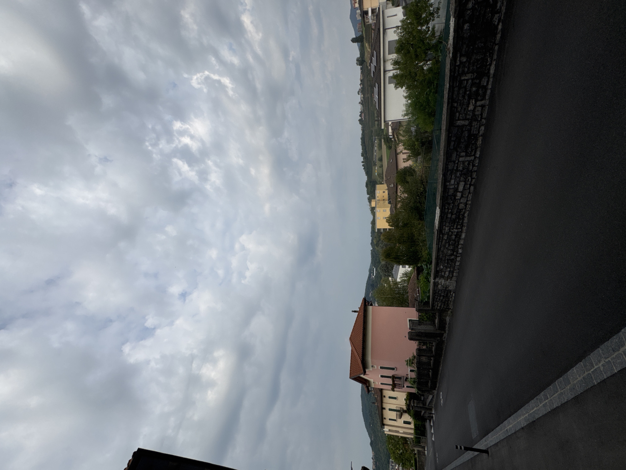 The image depicts a view of a street in Balerna, Switzerland, on a cloudy day. The foreground shows a dark asphalt road with a granite curb and a small black bollard on the left side. Beyond the road, a stone wall separates the street from a residential area. Several houses are visible, including a prominent pink building with a red tiled roof and a yellow building with multiple stories. In the background, rolling hills covered in vegetation and some terraced fields are present. The sky is filled with dramatic, grey clouds, suggesting overcast weather. The overall scene conveys a quiet, suburban or small-town atmosphere. There are no people or animals visible in the image.