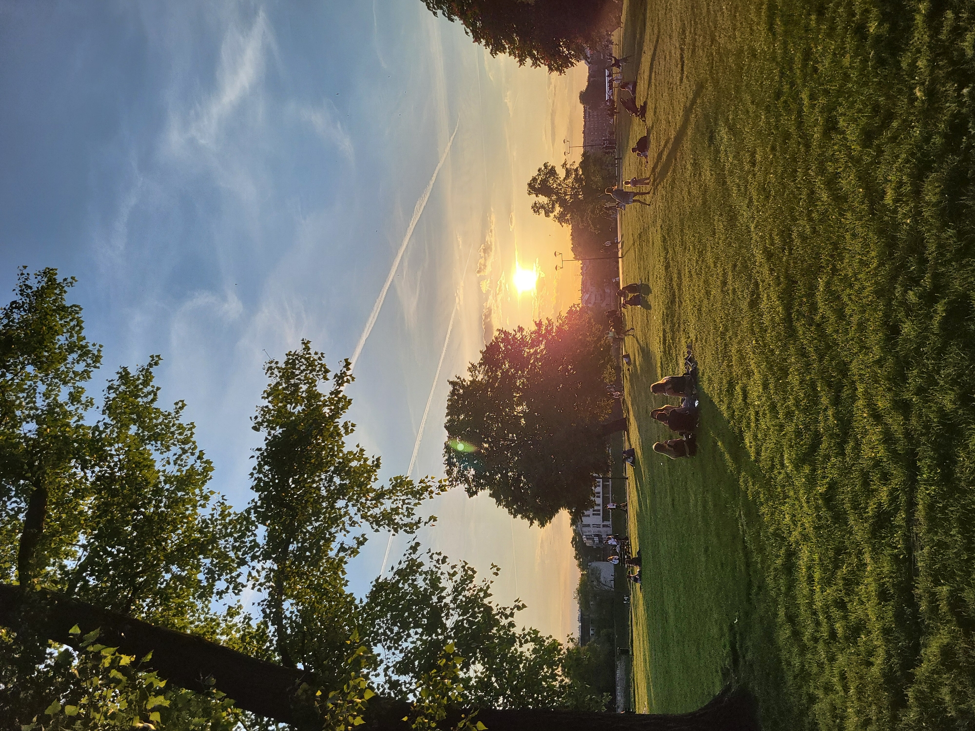 The image depicts a park scene at sunset. The sky is a gradient of blue to orange, with a visible contrail. Trees frame the view on the left and provide partial cover in the middle. The sun is positioned low in the horizon, casting a warm glow over the scene. People are scattered across the green lawn, some sitting in groups, others walking. The location appears to be in a city, evidenced by buildings in the distance.