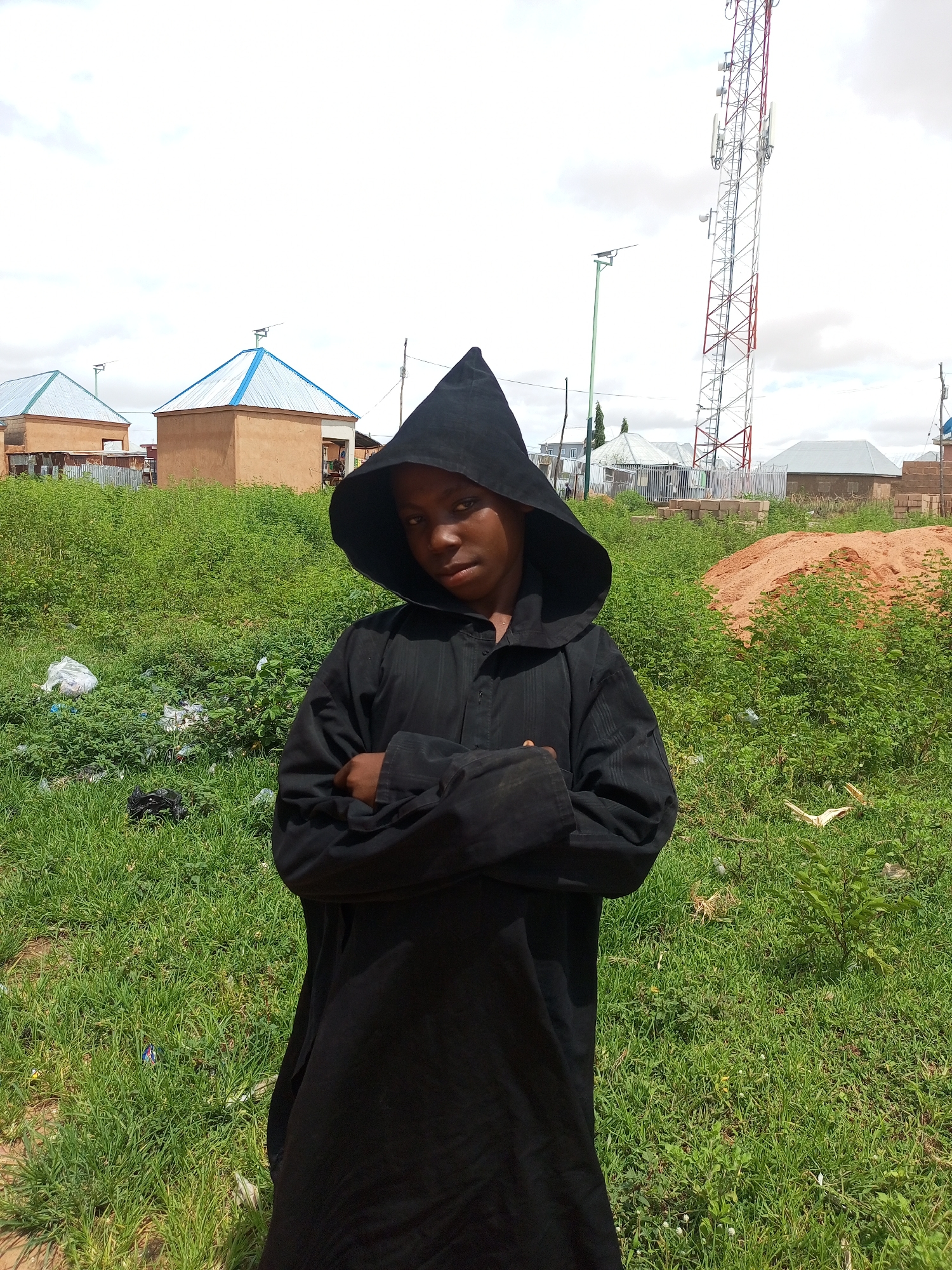 A person in a black hooded robe stands with arms crossed in a grassy, unpaved area. Behind them are several small, tan buildings with blue-trimmed roofs, a tall red and white telecommunications tower, and a pile of sand. The environment appears to be a developing area within the city of Tempure, Nigeria. The sky is overcast.