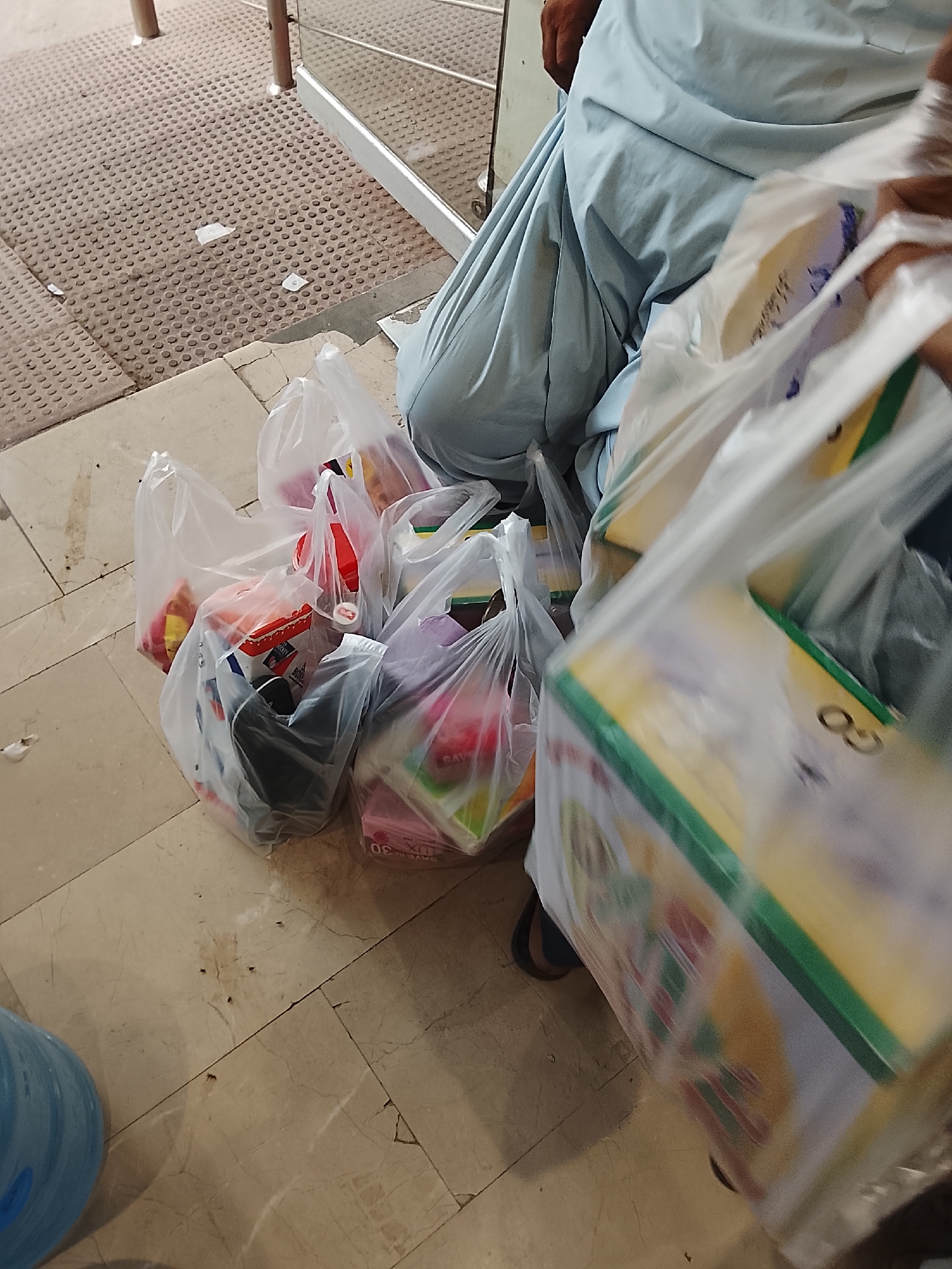 The image shows several plastic shopping bags filled with groceries on a tiled floor, likely inside or near a building in Gulgasht Colony, Pakistan. A person wearing light blue clothing is partially visible to the right, possibly the owner of the bags. The foreground is filled with blurred plastic bags, obscuring a clear view. The background shows a textured outdoor surface near a metal railing. The lighting appears to be indoor, artificial lighting. There are also some ants visible on the floor.