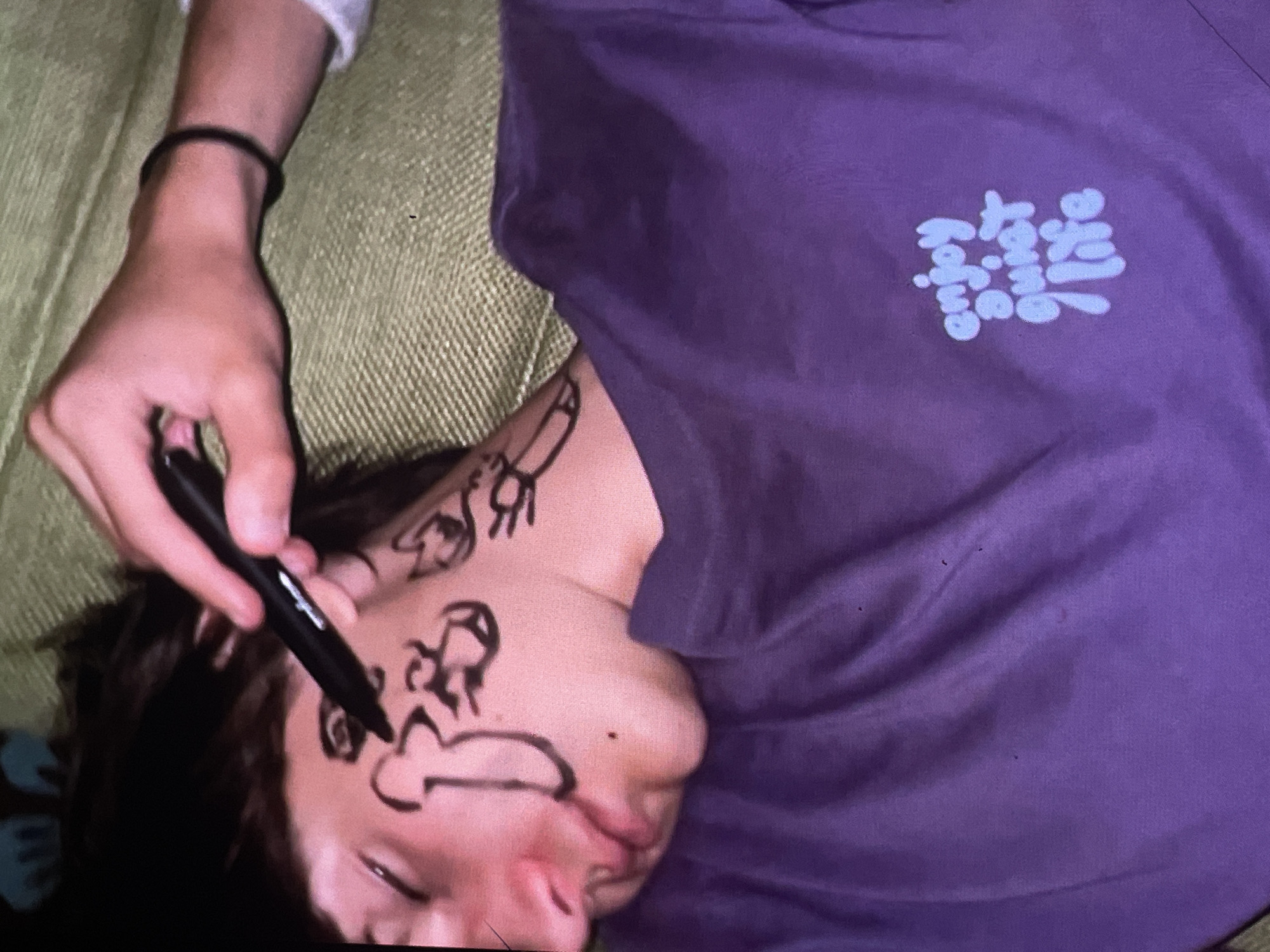 A person lies on a green textured surface, their face covered in black marker drawings. A hand is drawing on their face with a black marker. The person is wearing a purple t-shirt with "enjoy quiet life" written on it in a light blue font. The scene is indoors, and it is not possible to determine the city of Harmanli, Bulgaria.