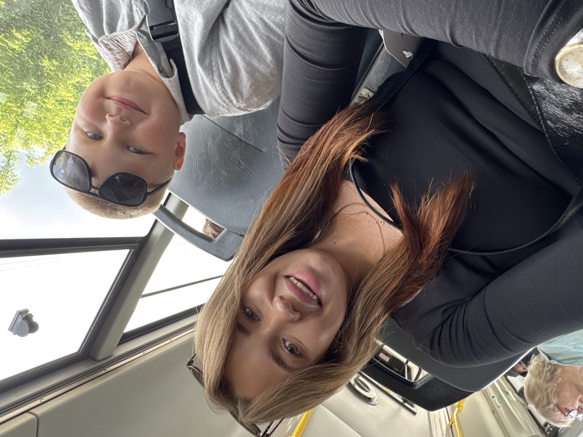 This is a photograph taken from inside a vehicle, possibly a bus or train, in Chaikovsky, Russia. The image is oriented sideways, with the subjects appearing upside down. The primary subjects are a young boy and a young woman, likely related or friends.
The boy is in the foreground on the left, wearing a light gray hooded sweatshirt and dark sunglasses. He is smiling and looking towards the camera. Behind him, out the window, green foliage and trees are visible, suggesting it is daytime and likely pleasant weather.
The young woman is on the right side of the frame, also looking at the camera and smiling. She has long, reddish-brown hair and is wearing a black top and black pants. She has a necklace with a small heart pendant visible. A watch with a gold face is also visible on her wrist.
The interior of the vehicle is visible, with dark gray seating. The overall mood of the photograph is happy and casual, likely a selfie taken during a journey. There are no visible signs or text in the image.