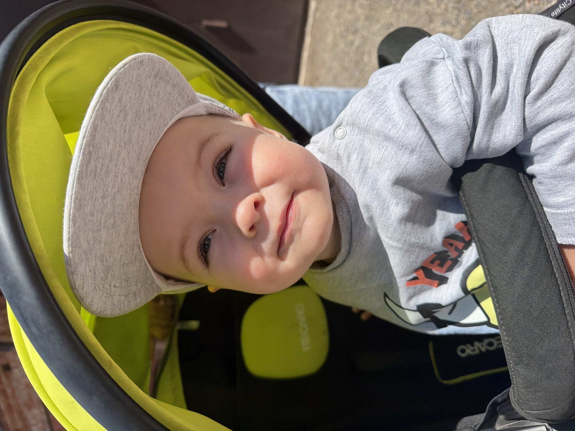 A baby is depicted seated in a Recaro stroller outdoors in Votkinsk, Russia. The infant, wearing a light gray baseball cap and a light gray long-sleeved shirt with a red "YEAH" graphic and a cartoon character, looks upwards with a slight smile. The stroller features a neon yellow/lime green canopy with a black frame, and the "RECARO" brand name is visible on a light green patch within the seat. A black harness or bumper bar is positioned across the baby's chest. Sunlight illuminates the scene, casting shadows. Partially visible in the background are a brick structure and a concrete ground surface. A person's leg, clad in light blue jeans, is visible to the right of the stroller. The word "Citylife" is subtly visible on a black element of the stroller near the baby's right arm.