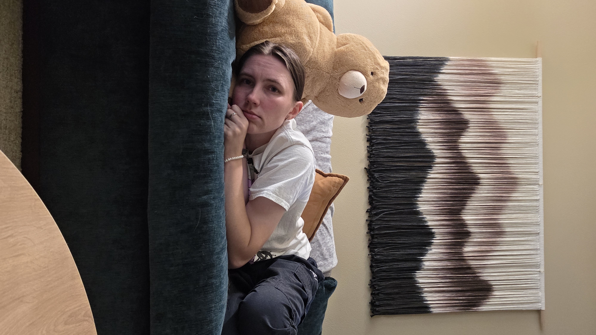 A young person is lying on a couch, tucked in with a large teddy bear. They are wearing a white t-shirt and dark pants. Their left hand is propped up near their chin, and they have a thoughtful or melancholic expression. A pearl bracelet is visible on their left wrist. A small orange pillow is behind them, and a large, plush teddy bear is positioned above their head.
The setting appears to be indoors, likely a living room, with a large, dark teal couch taking up the left side of the frame. To the right of the couch, a wall is visible, decorated with a large piece of textile art. This art features a gradient of colors, transitioning from black to white with shades of brown and pink in between, arranged in horizontal lines with a fringe at the bottom. A light-colored wooden table edge is visible in the bottom left corner. The lighting suggests it is daytime, possibly late afternoon or evening due to the warm tones on the wall. The overall mood is subdued and introspective.