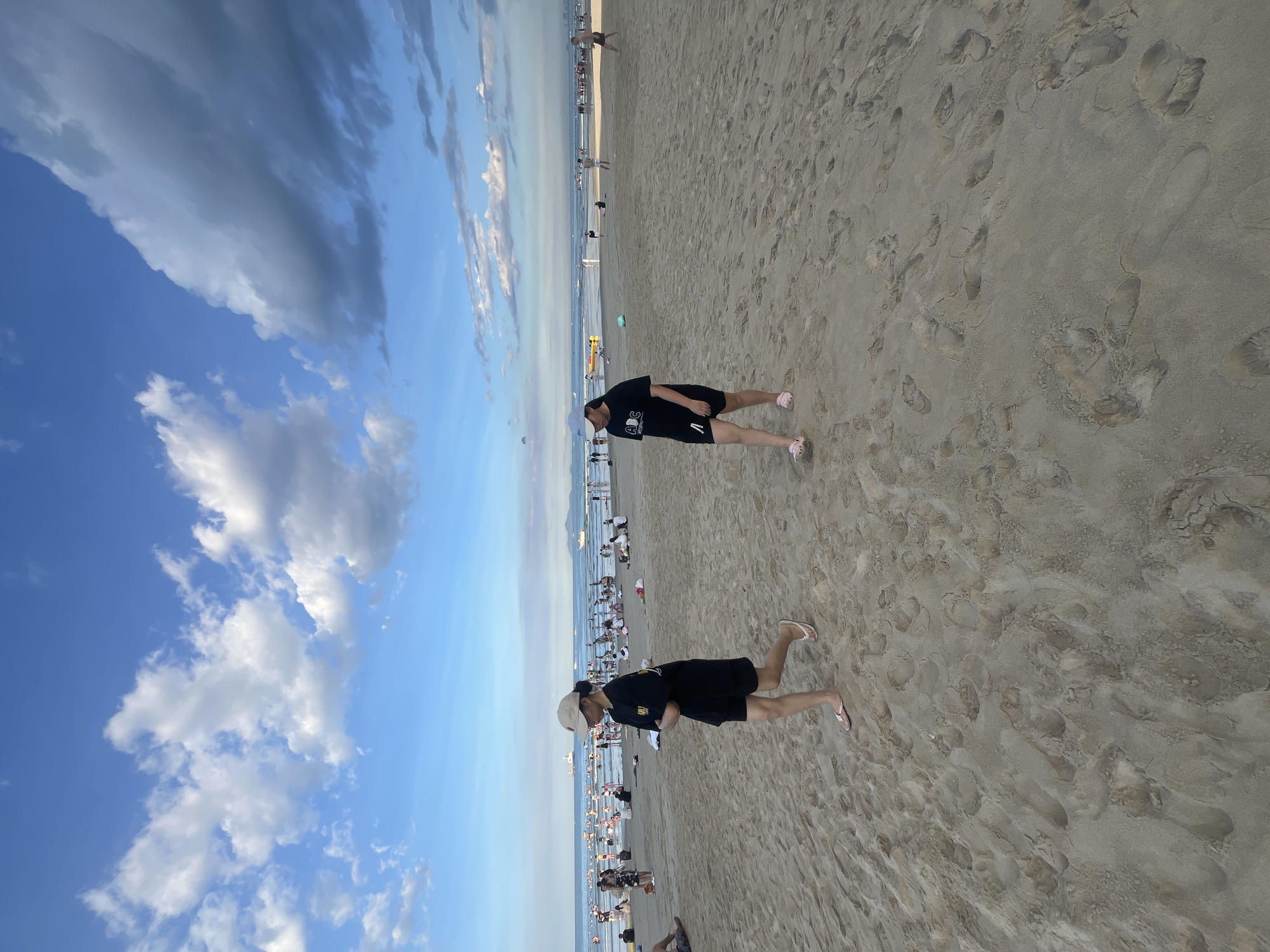 The image depicts a wide sandy beach in Đà Nẵng, Vietnam, under a partly cloudy sky. In the foreground, two young women are walking on the sand. The woman on the left is wearing a black t-shirt, black shorts, and white flip-flops, with a beige cap on her head. The woman on the right is wearing a black t-shirt, black shorts, and pink slides, also with a cap.
The beach is populated with numerous people further down, engaged in various activities. Some are sitting on the sand, others are walking towards the water, and a few are in the shallow sea. In the background, there are several boats and what appears to be a paraglider in the air. The ocean stretches to the horizon, meeting a hazy sky with several large clouds. The sand is covered with footprints, indicating significant foot traffic. The overall scene captures a lively beach atmosphere during the daytime.