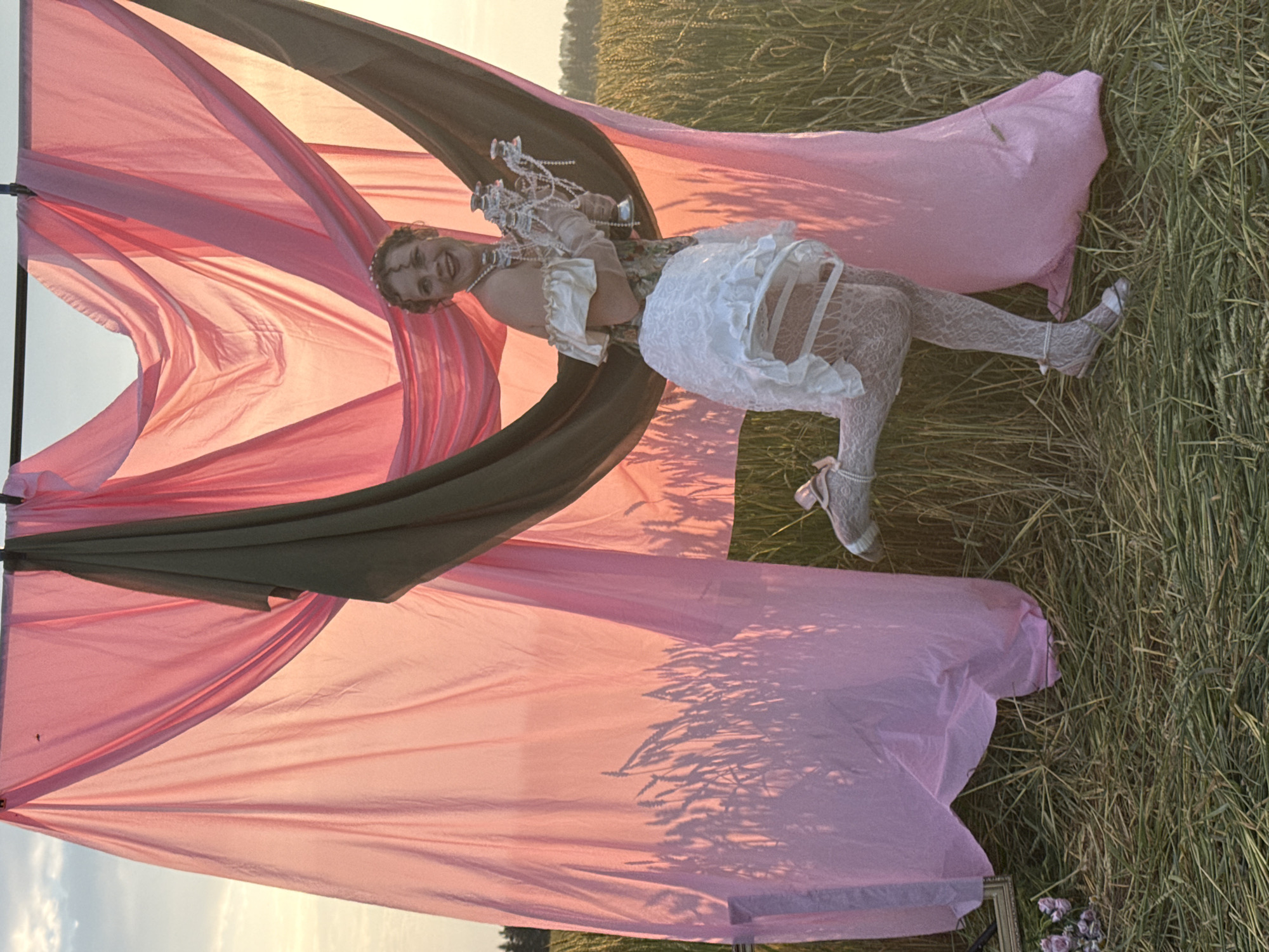 In Foki, Russia, a young person is positioned amidst a field of dry grass, partially enveloped by flowing pink and olive green fabric. The fabric creates a dramatic, draped effect, with sunlight casting shadows of the grass onto the pink material. The person, lying down, is adorned in a whimsical outfit. They wear white lace stockings with delicate ribbons, a frilly white skirt, and a pearl necklace. Their hair is styled with pearls as well, and they are holding a cluster of crystal-like decorations. Their expression is joyous and lively, with a wide smile. The scene is bathed in the warm glow of either sunrise or sunset, indicated by the soft, golden light and long shadows. The background suggests an open field, likely a meadow or agricultural land. The overall impression is one of artistic expression and playful elegance within a natural, rustic setting.