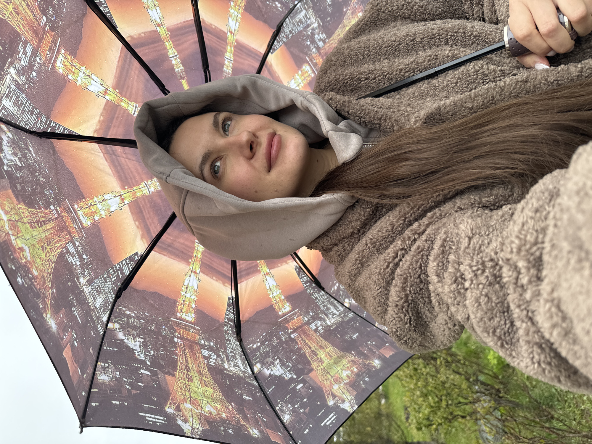 A woman with fair skin, light brown hair, and green eyes is holding an umbrella with an image of the Eiffel Tower and the city of Paris printed on it. She is wearing a light brown hooded sweatshirt and a furry brown jacket. The background shows a blurred outdoor scene with green trees. The location is unknown and cannot be determined as the image of the Eiffel Tower on the umbrella does not indicate that she is in the city of Odintsovo, Russia.