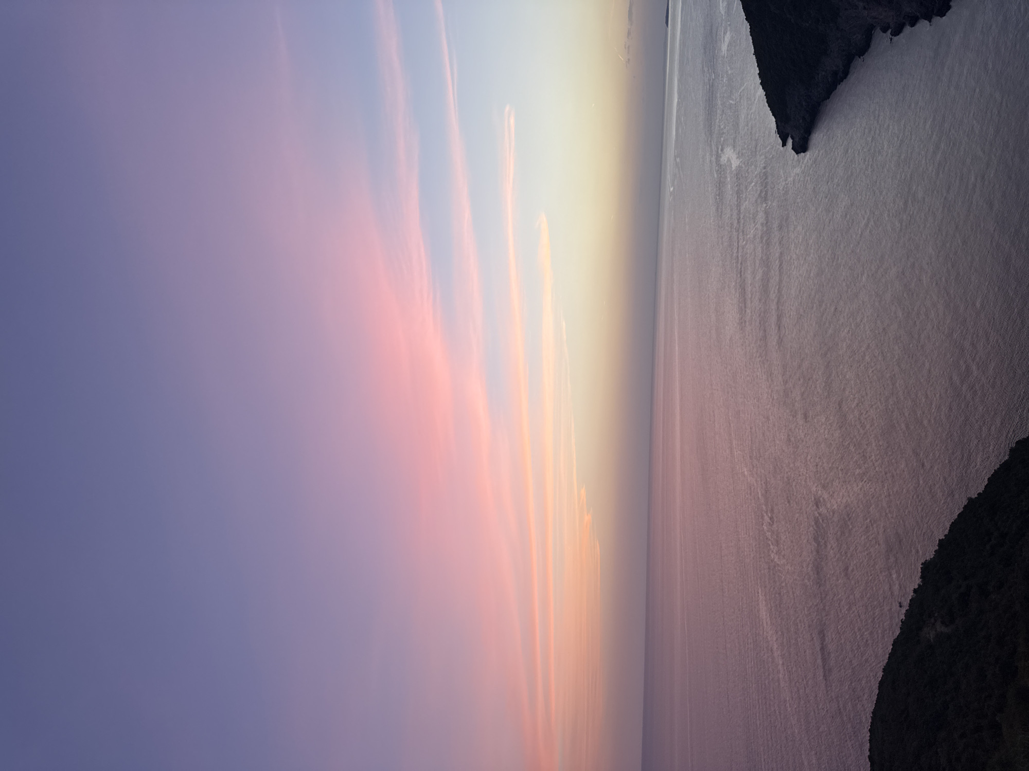 The image depicts a serene coastal landscape at dusk, featuring a vast expanse of calm water under a sky painted with soft hues of purple, pink, and pale yellow. Wispy clouds streak across the horizon, catching the fading sunlight. In the foreground, a dark, tree-covered hillside slopes down towards the sea on the left, while a rugged, dark coastline juts out into the water on the right, forming a bay. The overall scene is tranquil and evokes a sense of peaceful solitude. This view is characteristic of the coastal areas surrounding Zagora, Montenegro, where the Adriatic Sea meets the mountainous terrain. There are no people or specific man-made objects visible in the image.