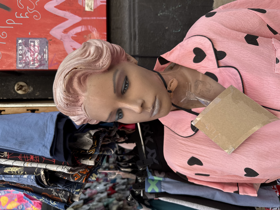 A mannequin with pink hair and wearing pink pajamas with black hearts rests on a pile of clothing. The mannequin's face is turned to the side, showing heavy makeup. The clothing pile consists of various colored shirts and pants. A red box with writing is visible in the background. The scene appears cluttered.