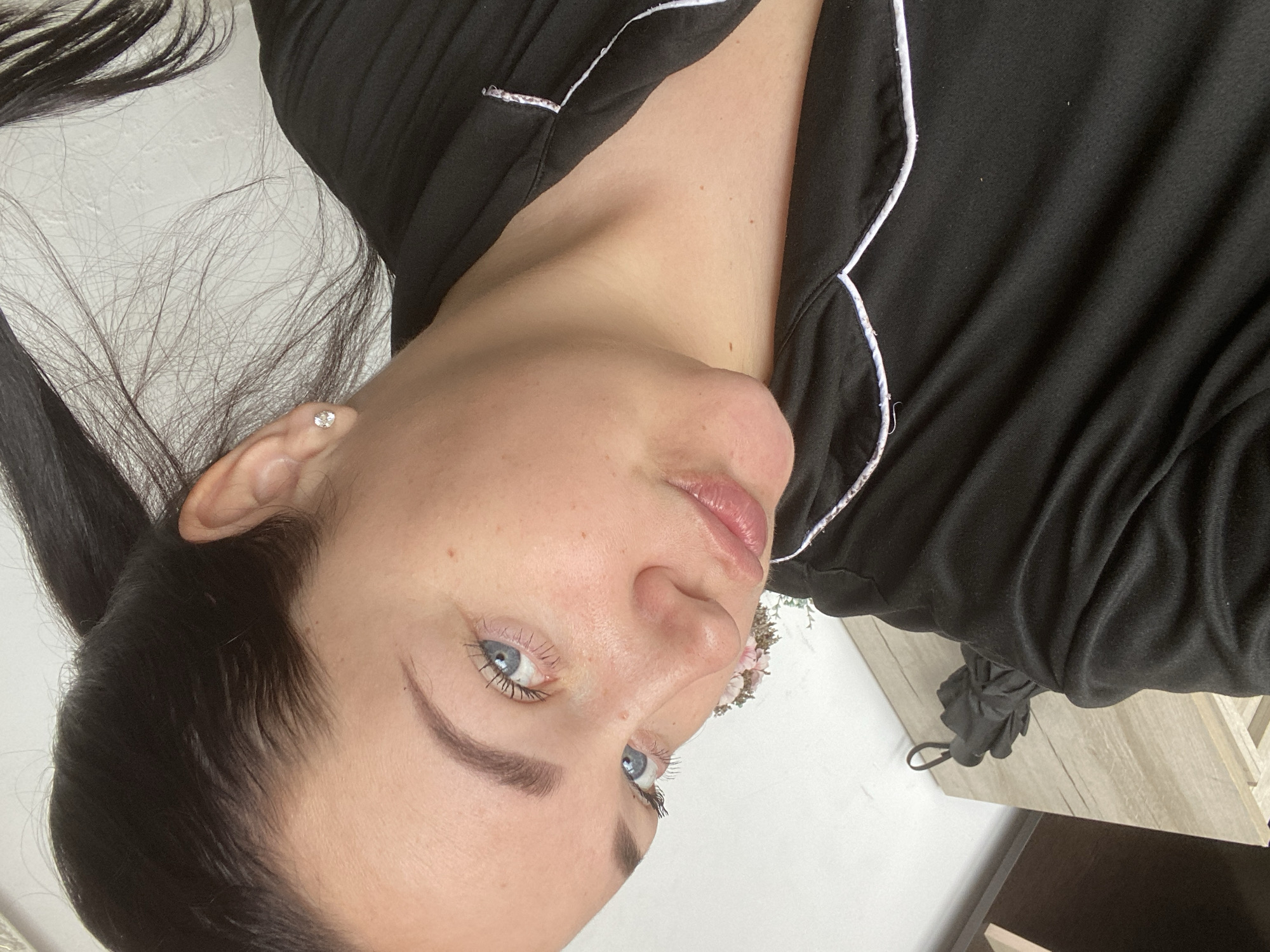 A portrait-style image, rotated 90 degrees, captures a young woman from the chest up, lying down and looking upwards. Her head is positioned on the left side of the frame. She has fair skin with visible freckles on her nose and cheeks, striking blue eyes with mascaraed lashes, and well-defined dark eyebrows. Her lips are slightly parted, showing a natural pink hue with a hint of gloss. Her dark hair is pulled back, with some fine strands visible around her face, and she wears a small, sparkling stud earring in her left ear. She is dressed in a casual black top, possibly pajamas or loungewear, featuring white piping along the collar and front opening. The garment's fabric shows a slight texture.
The background consists of light-colored wall or sheet to the left and a light wooden tabletop or nightstand to the right. A small, dark, bundled object with a loop, resembling a compact umbrella or a small bag, rests on the wooden surface. A hint of light pink and white floral decoration is visible near her head. The woman's expression is relaxed and calm. The scene depicts an intimate, indoor moment, likely in a home setting in Sochi, Russia. There is no visible text in the image.