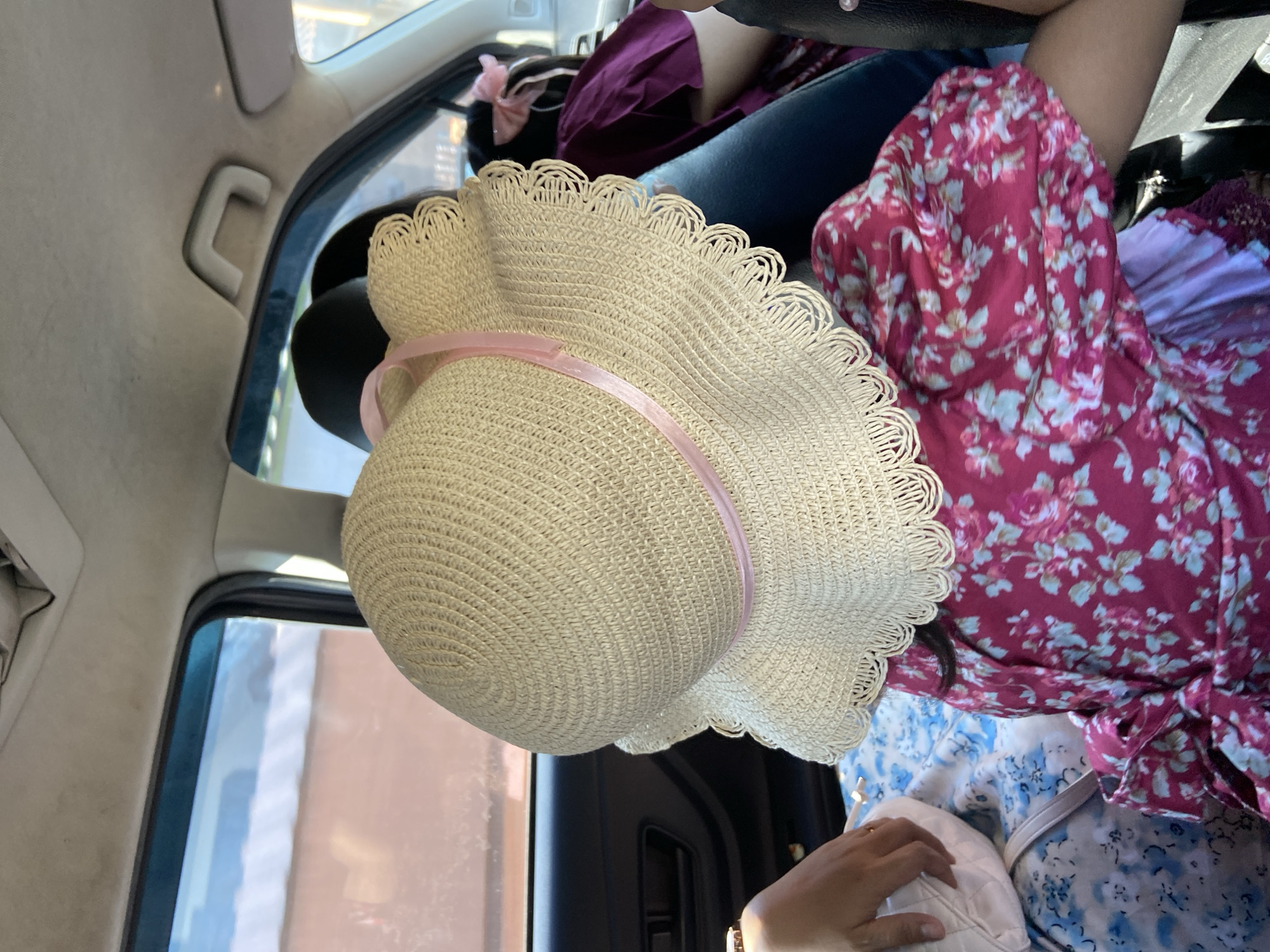 The image appears to be taken inside a car, possibly while in transit in Muktisari, Indonesia. The main subject is a light-colored straw sun hat with a pink ribbon, positioned prominently in the center. Parts of at least two individuals are visible, including a person wearing a dark floral print top and dark jeans. A second individual is wearing a blue floral dress, partially obscured. The person is holding a cream colored purse. The car interior details include a window, door frame, seat, and headrest. The environment is a car interior.
