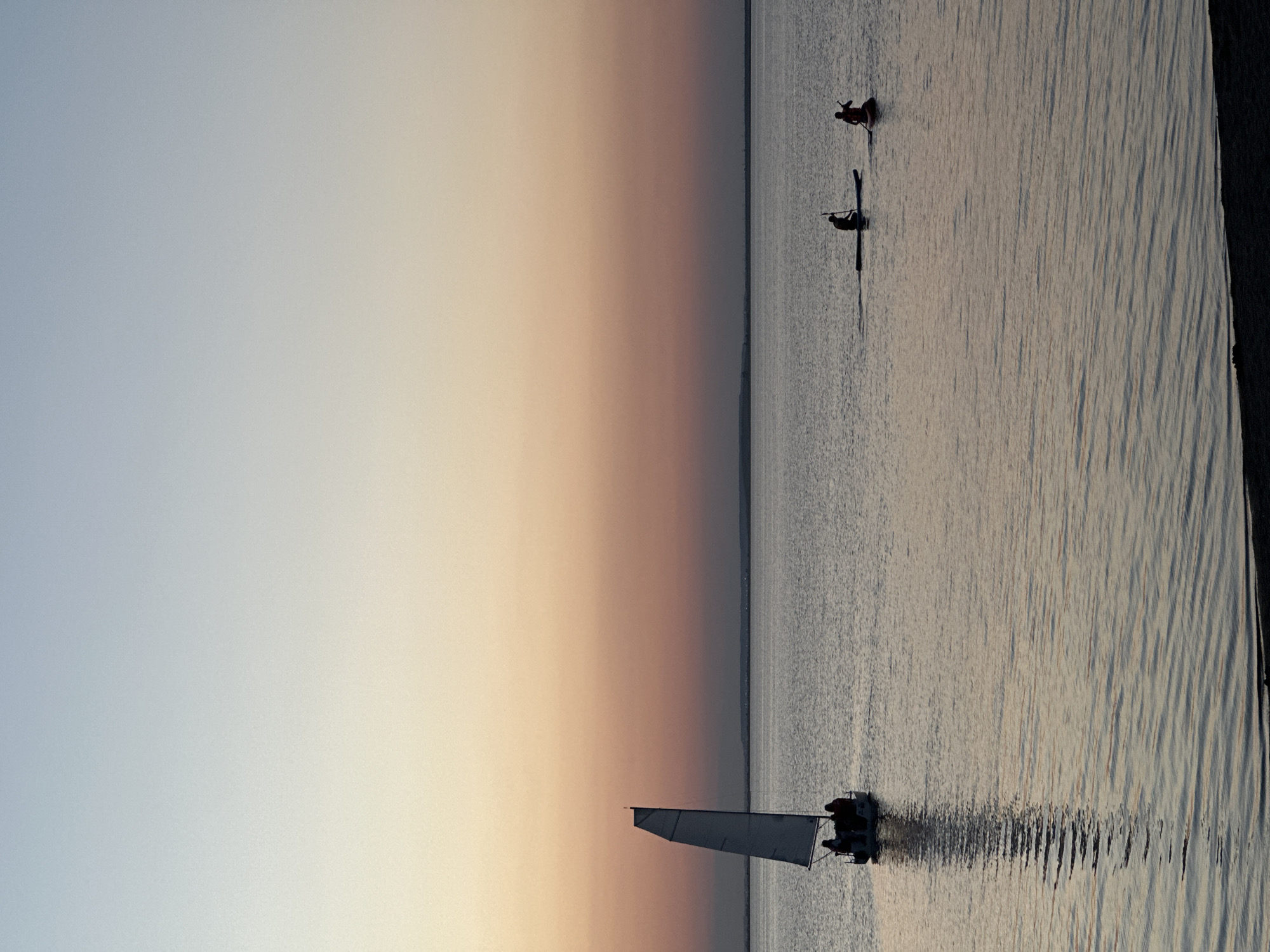 The image depicts a serene water scene at dusk, likely in Kokshetau, Kazakhstan. The sky transitions from a pale blue to a soft peach hue, indicating either sunrise or sunset. The calm water reflects the sky's colors and is rippled, suggesting a gentle breeze.
In the foreground, a small sailboat with a white sail is navigating the water. Two figures are visible on the boat, silhouetted against the fading light. Further in the distance, two individuals are on what appear to be kayaks or paddleboards, also silhouetted. They are positioned further out on the water, and their dark forms create a stark contrast with the lighter water.
The overall mood is tranquil and peaceful, capturing a quiet moment on the water as day transitions to night. The distant horizon line is barely visible, blending into the sky, creating a sense of vastness. There is no visible text in the image. The lighting and the activity suggest a leisurely pursuit during a calm evening or early morning.